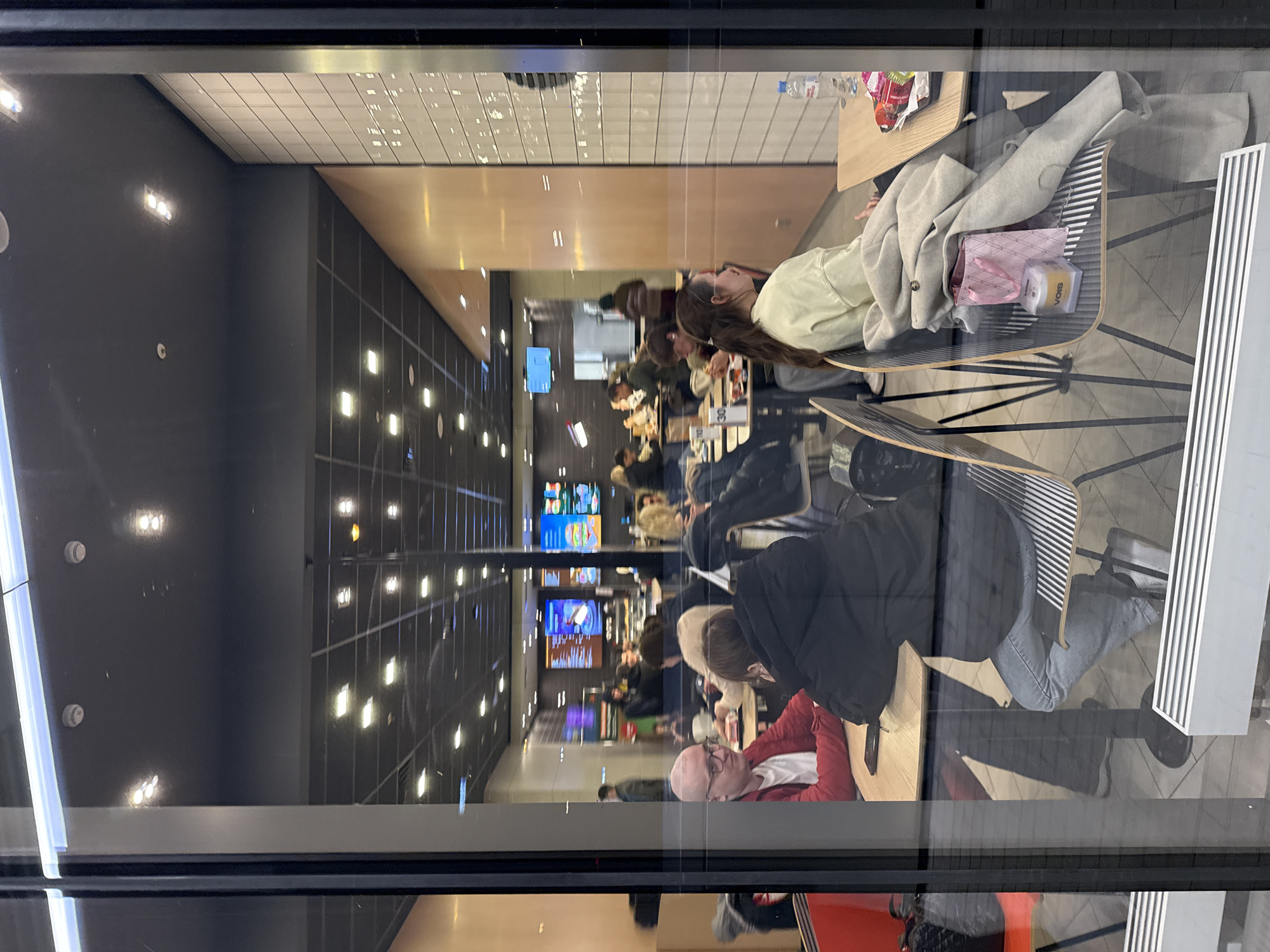 The image shows the interior of a brightly lit, modern fast-food restaurant or cafeteria, viewed through a large glass window. The photograph is oriented vertically, rotated 90 degrees counter-clockwise.
Numerous people are seated at light-colored wooden tables, engaged in dining or socializing. In the mid-right of the frame (when viewed correctly), a woman with long, dark hair, wearing a light yellow top, sits at a table. Next to her, a light gray jacket is draped over a chair, alongside a small pink bag and a clear plastic bottle with the brand name "VOIS" clearly visible. A small table stand displaying the number "30" is also present. Further into the restaurant, a bald man in a red top is visible among other diners.
The restaurant features a contemporary design with dark gray walls and a ceiling composed of both dark panels and white tiled sections, illuminated by multiple bright, rectangular light fixtures. In the background, several digital menu screens, characteristic of fast-food establishments, display various items and advertisements. Reflections are visible on the glass window through which the image was taken, showing elements of the outside environment.
The scene depicts a casual mealtime setting in Khimki, Russia. The interior lighting suggests it could be day or evening. The people appear relaxed, with no discernible strong emotions.