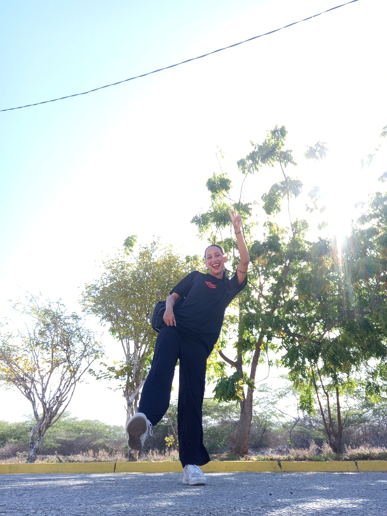 A full-length image shows a person standing on one leg on an asphalt surface with trees and foliage in the background. The person is wearing a dark-colored T-shirt and dark pants, holding one arm up in the air with two fingers extended. They appear to be smiling. The ground is paved and marked with a yellow curb. The background features trees and other foliage, with bright sunlight filtering through the branches. A single wire is visible at the top of the image against the bright sky. The location appears to be in a natural, outdoor setting within the city of Porlamar, Venezuela.