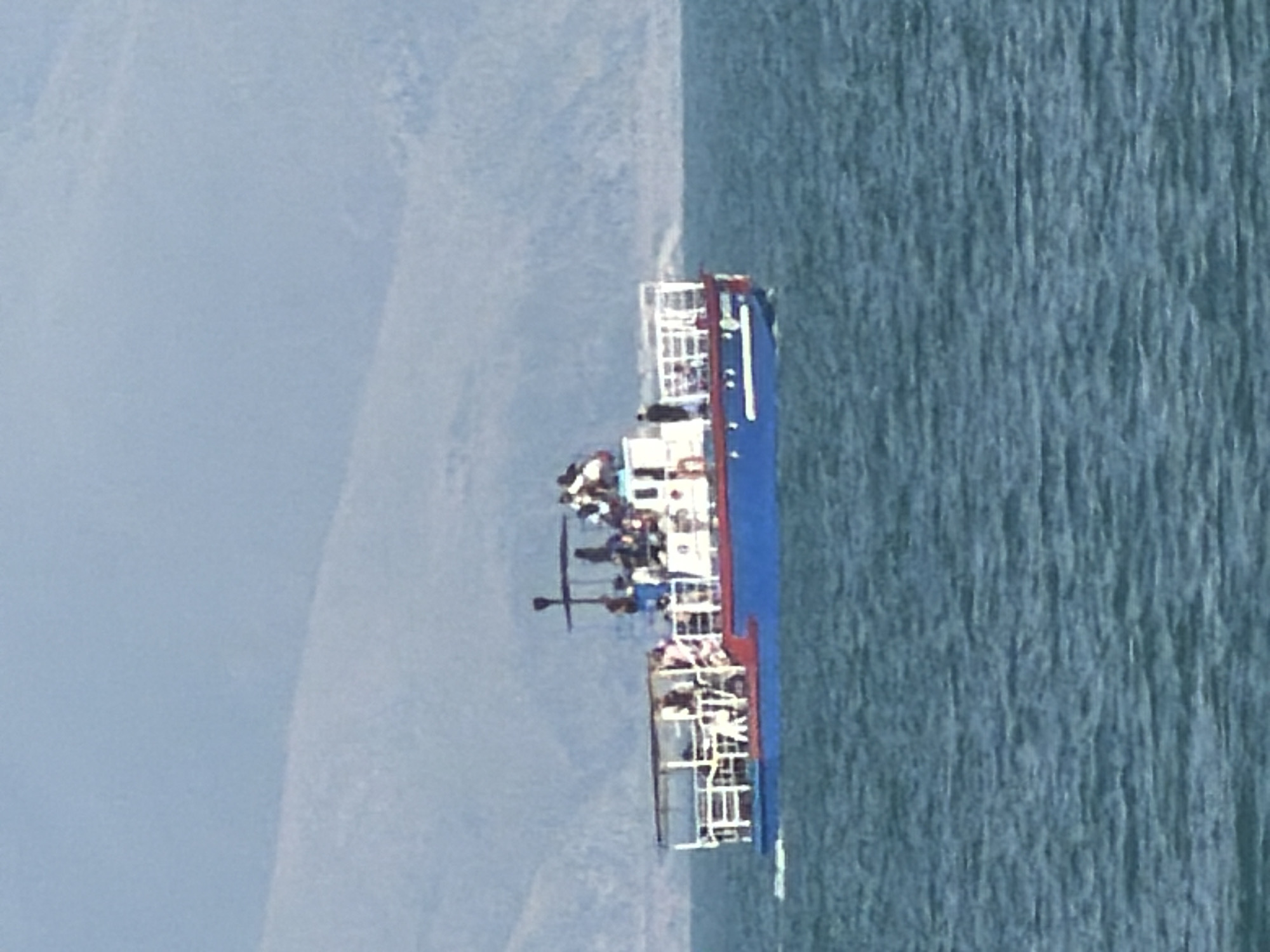 A blue and white ferry boat with a red hull is sailing on a body of water, likely a lake, with a faint, misty mountain range in the background. The ferry is populated with many people, some standing on the upper deck and some on the lower deck, which is partially covered by a white canopy. The water is a deep blue-green with small ripples. The sky is a pale, hazy blue. The scene appears to be a picturesque day for a boat trip. While Tsamakaberd is in Armenia, this image does not contain specific landmarks to confirm its location.