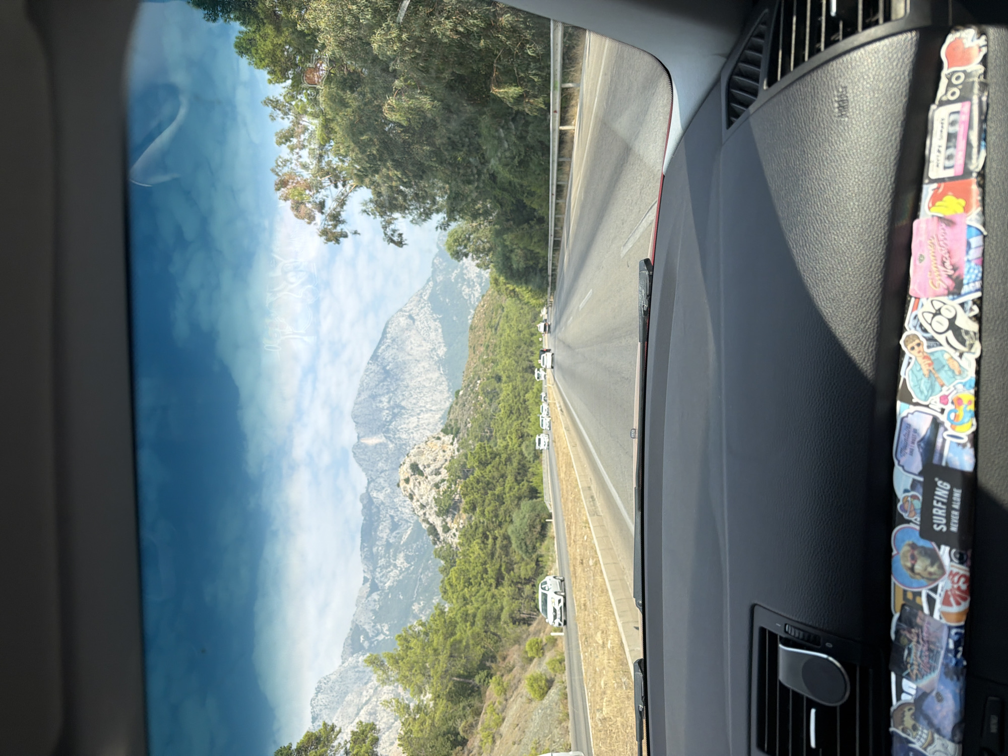 The image shows a view from the driver's seat of a car on a highway. The car's dashboard is visible in the foreground, adorned with numerous stickers. The road ahead is lined with trees and mountains, suggesting a scenic route. Several cars are driving on the highway. The sky is blue with scattered clouds. The location is identified as Kemer, Turkey.