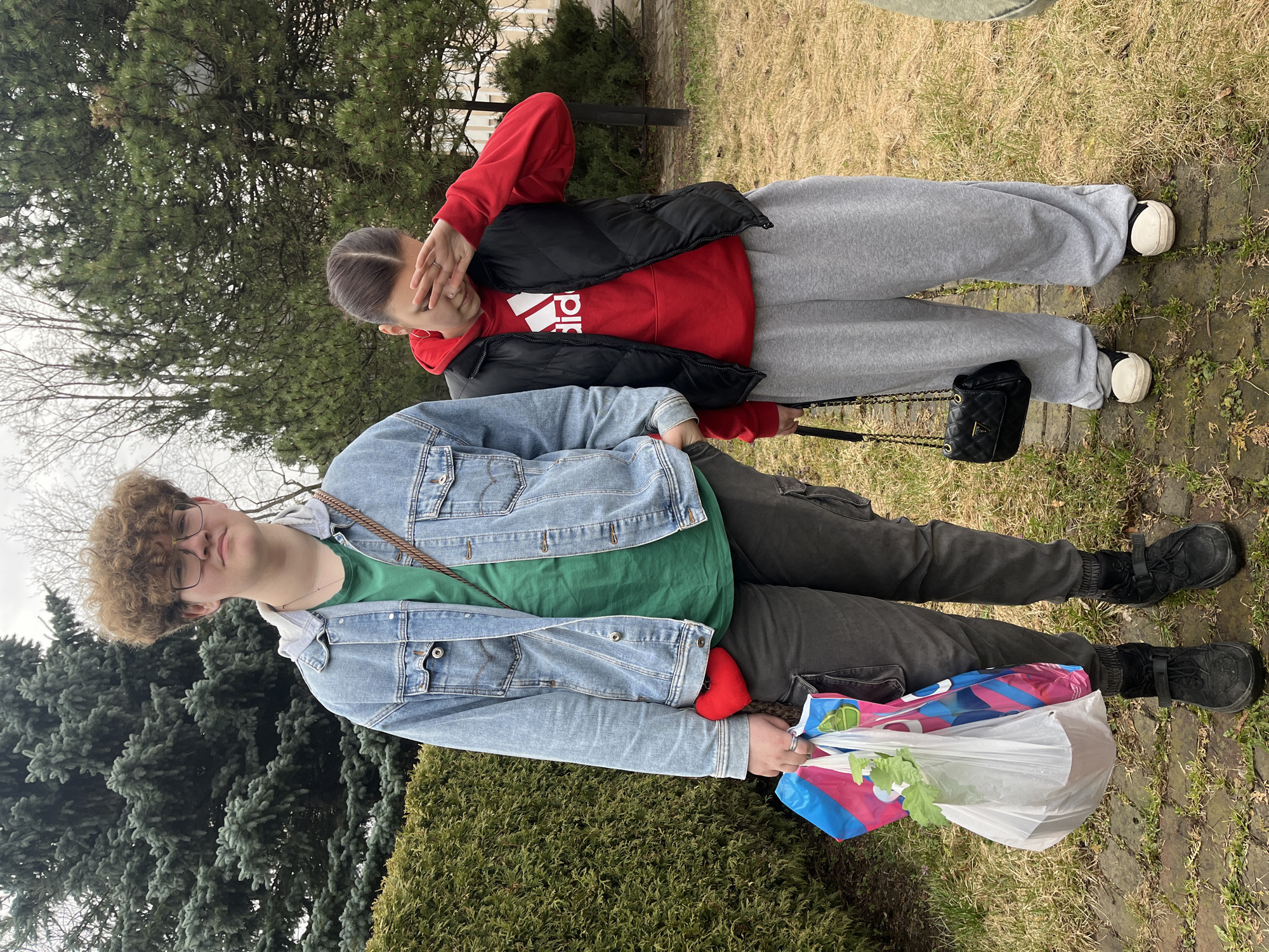 Two young people are standing outdoors. The person in the foreground has curly blonde hair and wears glasses, a green t-shirt, a blue denim jacket, dark pants, and black shoes. They hold a plastic bag. A red heart-shaped object is tucked under their arm. Behind them, the second person wears a red hoodie with a black vest, grey sweatpants, and black and white shoes. They are obscuring their face with their hand. A black purse hangs from a chain over their shoulder. The location appears to be outdoors, possibly a park or garden, given the presence of trees, bushes, and grass. Without any additional information or context, it is not possible to confirm if the image was taken in Saint Petersburg, Russia, or in any other location.