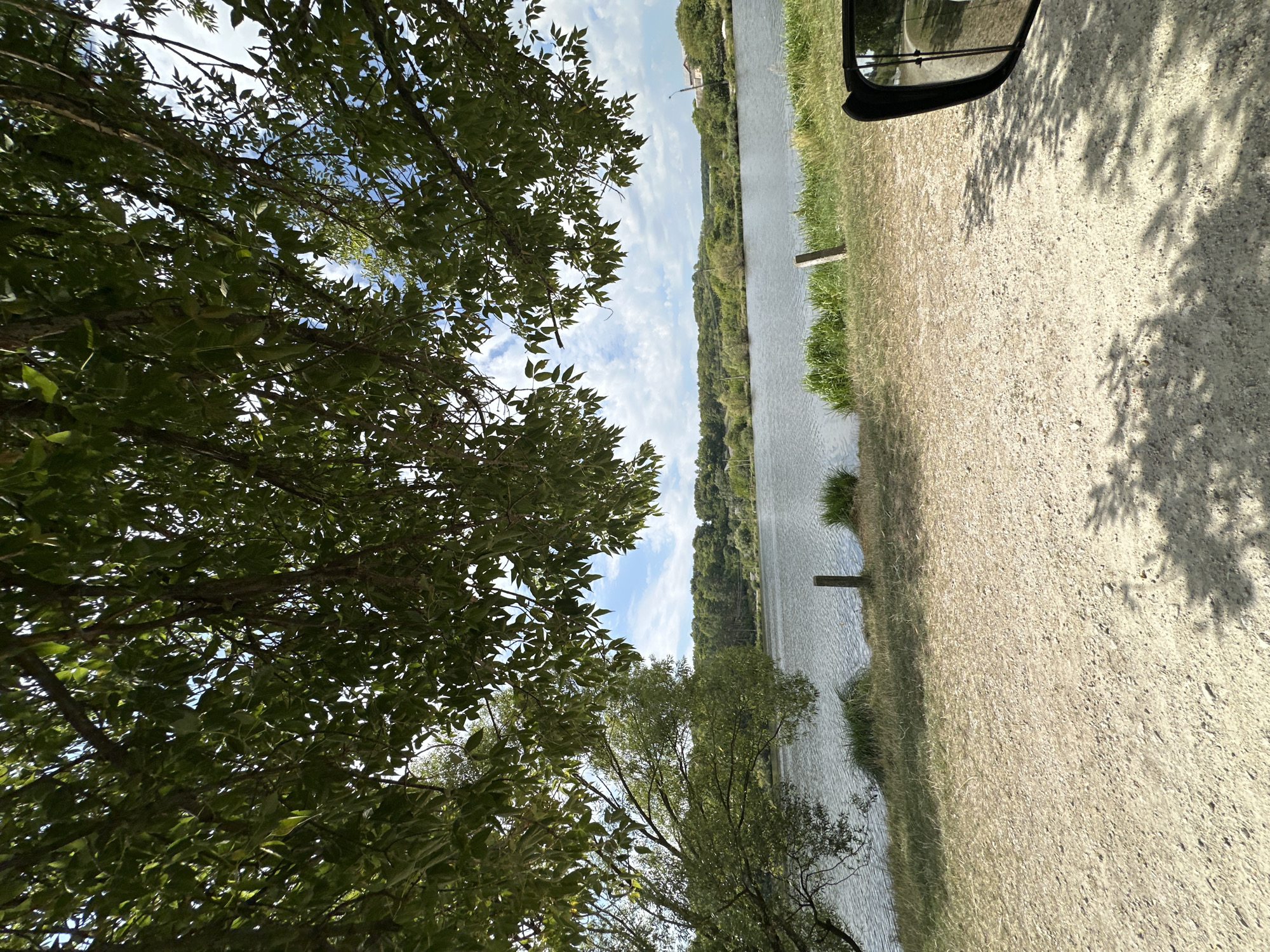 The image depicts a serene natural landscape, likely a lake or riverfront, as seen from a slightly elevated, sandy vantage point. Overhanging green foliage from trees frames the top of the image, dappled with sunlight. In the middle ground, a body of water stretches across the frame, its surface rippled by a gentle breeze. The far bank of the water is lined with dense green trees and some scattered buildings, suggesting a rural or semi-rural setting. The foreground consists of a light-colored, gravelly ground, with shadows from the trees cast upon it, indicating a sunny day. A portion of a dark-colored vehicle's side mirror is visible on the right edge of the frame, suggesting the photo was taken from inside or next to a vehicle. The sky above is blue with scattered white clouds. Specific to Makiv, Ukraine, this scene evokes a peaceful, natural environment that could be characteristic of the area, known for its natural beauty.