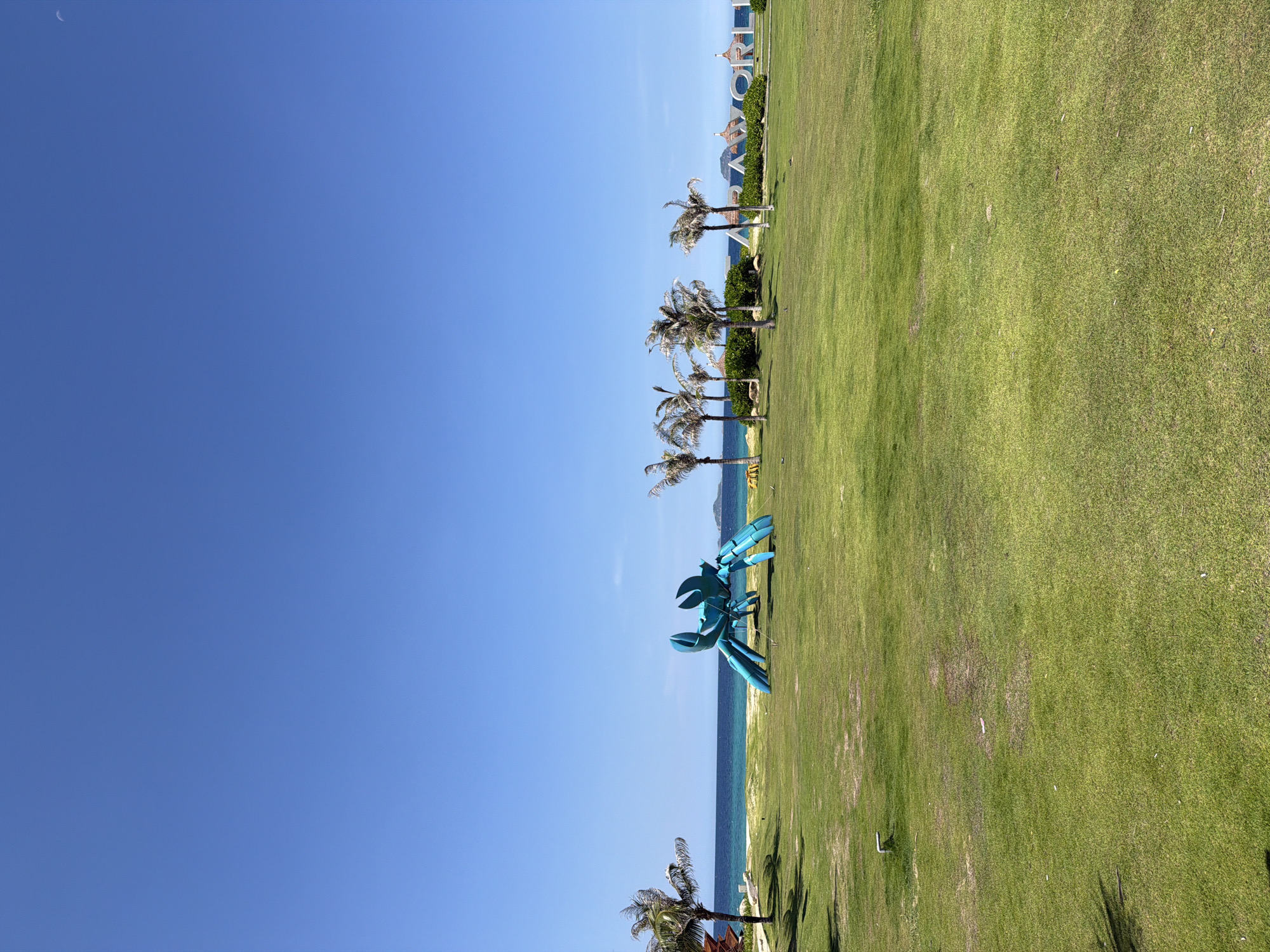 The image shows a bright, sunny day at a beach resort. The foreground is a manicured green lawn with a few scattered white objects. In the mid-ground, there is a large, bright blue sculpture of a crab. To the right of the crab, there are several palm trees and in the far background, a sign that appears to read "WORLDC" in large white letters, with some thatched roof structures. The ocean is visible beyond the lawn, with a clear blue sky above. The location is identified as Bac Cam Ranh Ward, Vietnam. There are no people or animals visible in the image.