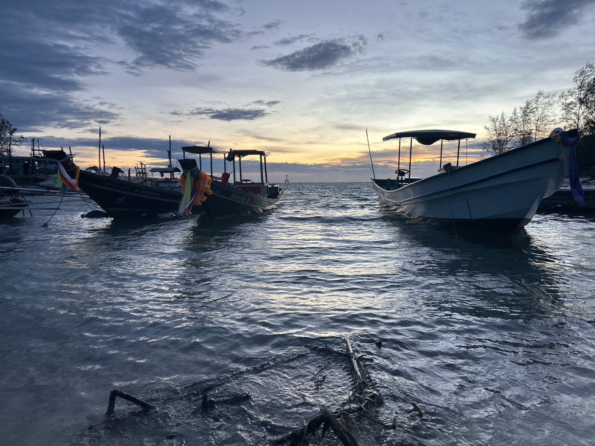 The photograph captures a seascape at dusk, with several small boats moored near the shore in Baan Sri Thanu, Thailand. The sky is a mix of grays, blues, and orange hues, indicative of either a sunrise or sunset. Three boats are prominent: two traditional Thai boats with overhead canopies are adorned with colorful garlands. The third boat, a light blue vessel with a similar canopy, is positioned to the right. The water is gently rippling and reflecting the sky's colors. Debris, possibly chains or metal structures, lies partially submerged in the foreground. No people are visible in the scene.