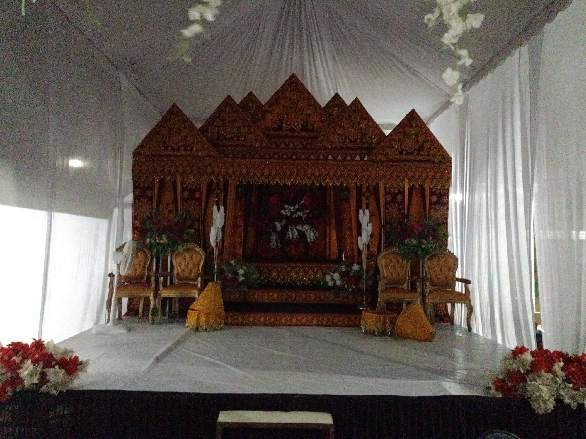A wedding stage is set up indoors in Seuneuboklhong, Indonesia. The stage is elevated and draped in white fabric, with flower arrangements on the front corners. A decorative, dark wood structure serves as a backdrop, featuring intricate patterns and multiple gabled roof-like shapes. Two ornate, upholstered chairs are placed on either side of the stage, with matching decorative items beside them. White drapes line the sides of the stage area.