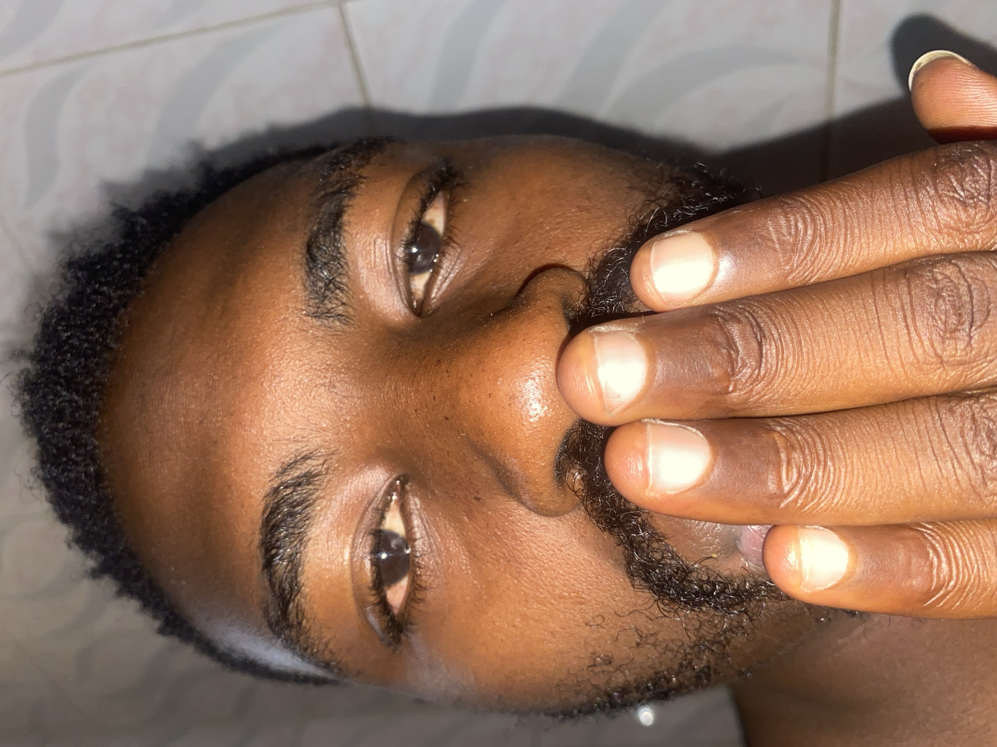 This close-up portrait shows a person of dark skin tone, with the image oriented sideways. The subject's face fills the frame, with their right hand positioned to cover their mouth and part of their nose.
The person has dark, curly hair, visible along the top and side of their head. They have dark eyebrows and stubble forming a beard and mustache. Their eyes are open and looking directly towards the viewer, reflecting some light. The skin on their face appears somewhat shiny, possibly from oil or sweat, under the direct lighting.
The hand covering the lower face has visible fingers and fingernails, which appear clean and of a natural, light color. The texture of the skin on the fingers shows natural creases and lines.
The background is a light-colored tiled wall, creating a simple and unobtrusive setting. Strong shadows are cast from the hand onto the face and from the head onto the wall behind, indicating a concentrated light source. The overall impression is an intimate, perhaps casual, indoor shot. There is no visible text in the image.