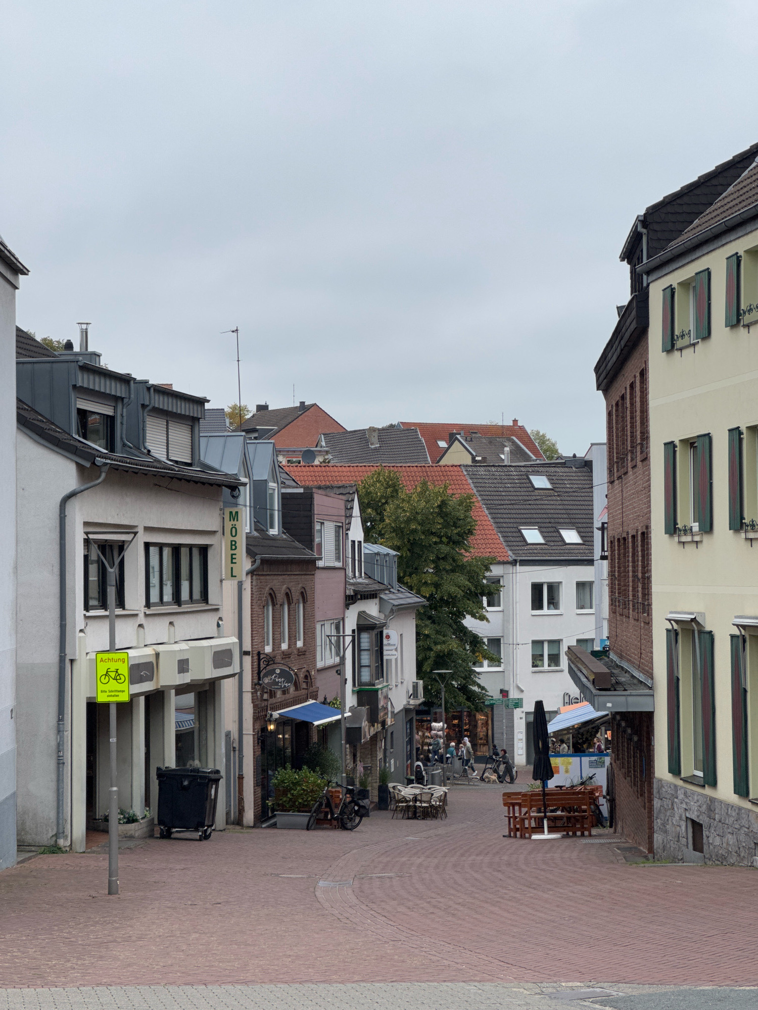 This is an eye-level, medium shot of a street in the city of Cleves, Germany, on an overcast day. The street is paved with red brick, and on either side are multi-story buildings with commercial establishments on the ground floor. The buildings display a mix of architectural styles, with some featuring traditional brickwork and others a more modern facade.
On the left side of the street, a building has a sign that reads "MÖBEL", indicating a furniture store. Further down the street, a yellow sign with a bicycle symbol and the word "Achtung" is mounted on a lamppost. A large black dumpster is situated near the entrance of the furniture store.
On the right side, a building with cream-colored walls has several windows with green shutters. A brick building with rows of windows is adjacent to it. Further down, a cafe or restaurant with outdoor seating is visible. People are seen walking and sitting at outdoor tables, suggesting a lively commercial area. Several bicycles are parked along the street. The sky is a uniform gray, indicating a cloudy day. Trees with green foliage are visible between the buildings.