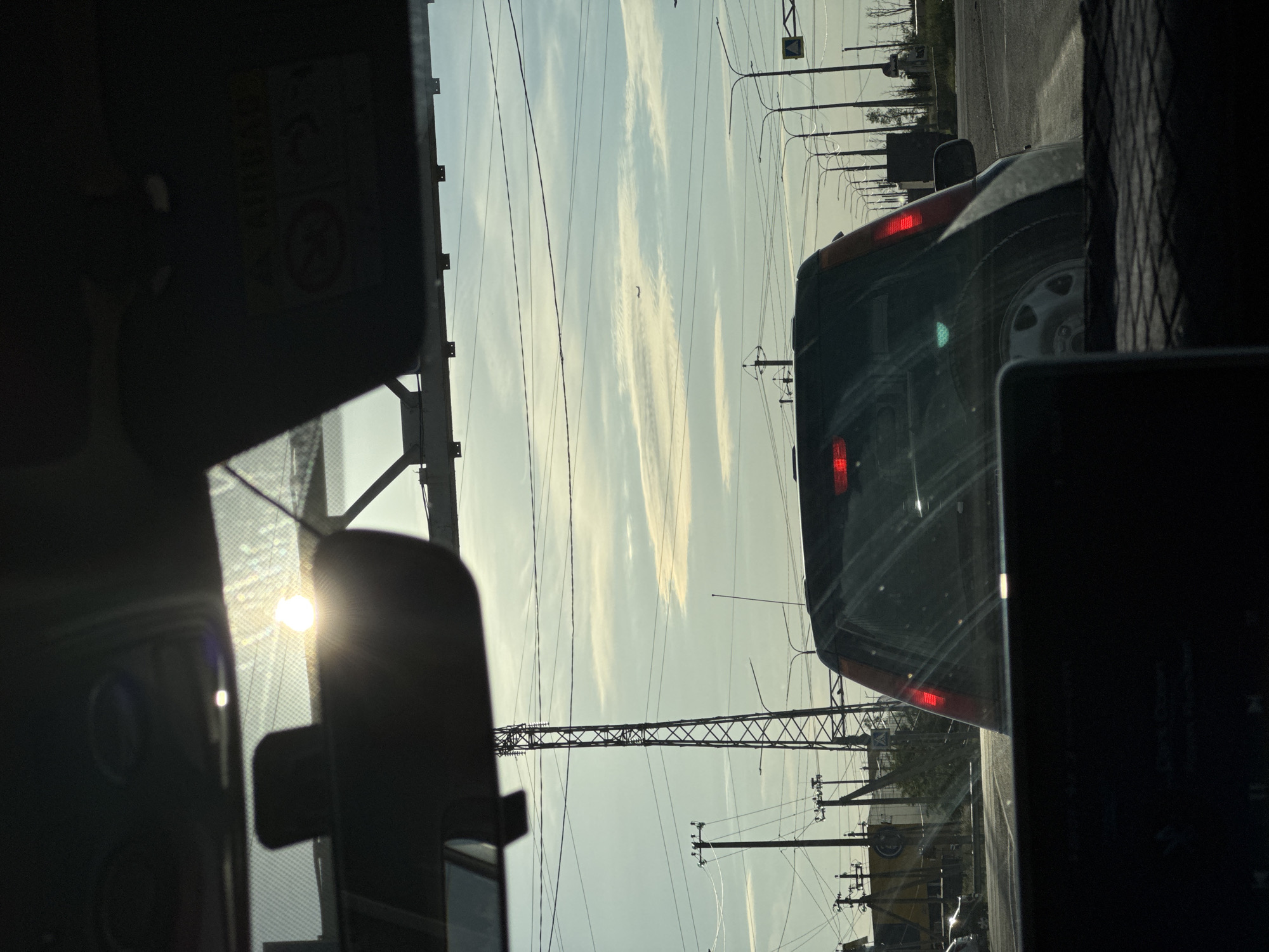 This media file is a photograph taken from inside a vehicle, looking out at a road scene. The perspective suggests the photographer is in the passenger seat, with the windshield and dashboard in the foreground. The sun is visible, partially obscured by the vehicle's interior, indicating it is either sunrise or sunset. The sky is a pale blue with wispy white clouds, suggesting fair weather.
The primary subjects are the road and other vehicles. A dark-colored SUV is in front of the photographer's vehicle, with its brake lights illuminated, suggesting it is either stopped or slowing down. There are numerous utility poles and power lines crisscrossing the sky, common in many urban and suburban areas. The landscape appears somewhat arid with sparse vegetation visible in the distance.
The setting is a road, likely a highway or a major road, given the presence of multiple lanes and the overhead power infrastructure. The presence of power lines and utility poles are strong location cues. The image depicts a common travel or commuting scene. There are no people clearly visible.
Notable details include the glare of the sun on the windshield, the red brake lights of the SUV, and the intricate network of power lines. There is some text visible on the side of the SUV in the distance that appears to be a logo or advertisement, but it is too blurry to read. A yellow sign with a black symbol is also visible on a pole. The overall mood is neutral, capturing a mundane moment of travel.
The file appears to be oriented vertically, suggesting it might have been captured by holding a phone in portrait mode or by rotating the camera. The location context of Novomar'evskaya, Russia, implies a setting in that geographical area.