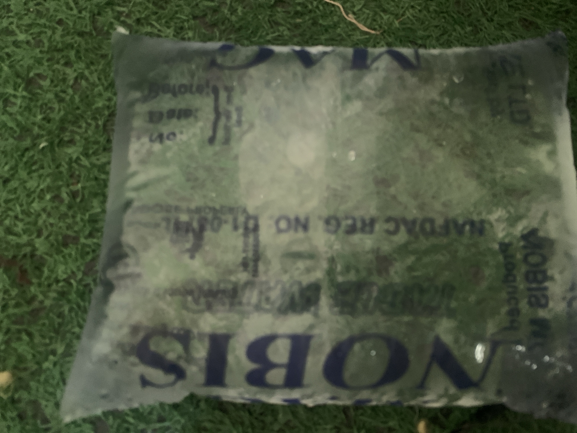 A clear plastic sachet, containing a green liquid, lies on a bed of artificial green turf. The sachet is rectangular and appears to be sealed at the top. Text is visible on the sachet, with the most prominent being the word "NOBIS" written vertically in large, dark blue letters. Below this, smaller text is present, including "NAFDAC REG. NO. 01-04". There are also some reflections on the surface of the sachet, obscuring some of the text. The image is taken from a slightly elevated angle, looking down at the sachet. The background is out of focus but appears to be a continuation of the green turf. The lighting is dim, and there is some glare on the plastic. The setting is not specifically identifiable as Jauro Yinu, Nigeria, and the image lacks any contextual clues to place it in that specific location.