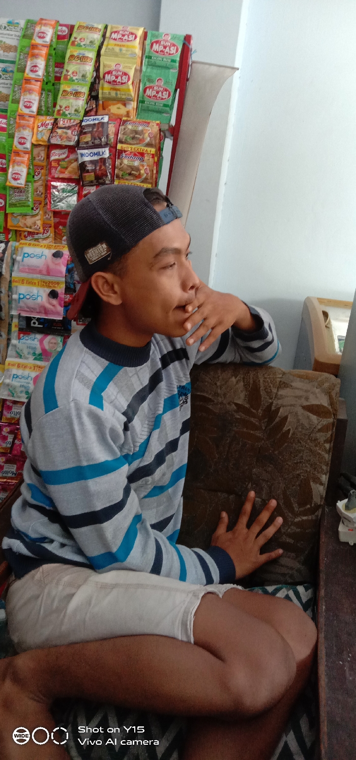 A young man in a striped shirt and baseball cap sits on a patterned cushion. He has one hand near his mouth and the other resting on the cushion. Behind him is a wall display of various food and household product packets. "MP-ASI" is visible on several packages. The bottom of the image includes text indicating it was taken with a Vivo Y15 AI camera.