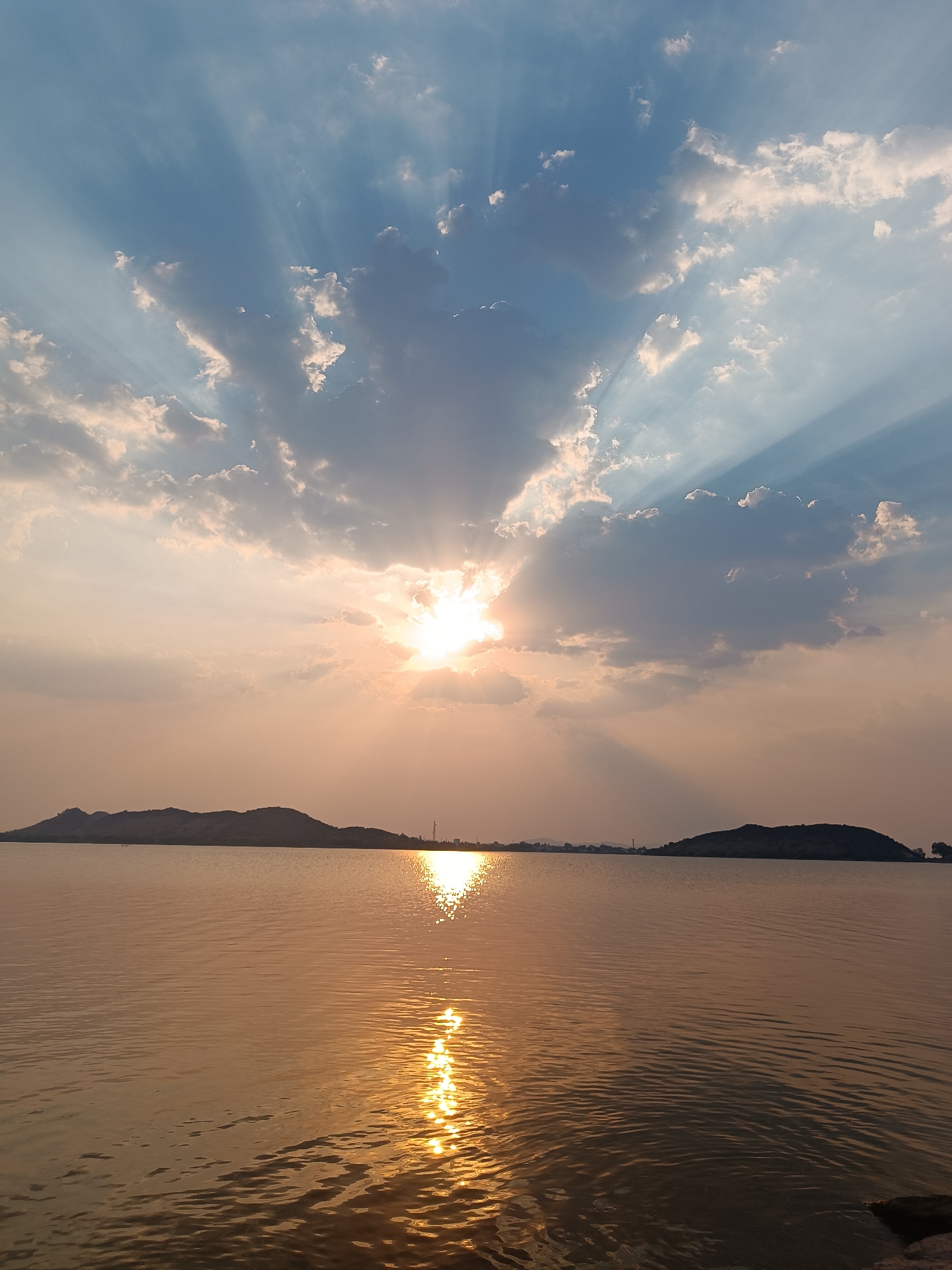 The image shows a wide body of water with two hills or small mountains visible in the distance under a sky with clouds and the sun. The sun is partially obscured by the clouds, but its rays are visible, especially above and around the clouds. The sunlight is reflecting off the water's surface, creating a bright, vertical path of light that extends from the sun's position to the foreground. The water appears calm with subtle ripples. The overall color palette is dominated by warm hues, particularly golds and yellows, due to the sunset or sunrise. The image appears to be taken at dusk or dawn, where the ambient lighting is soft and diffused.