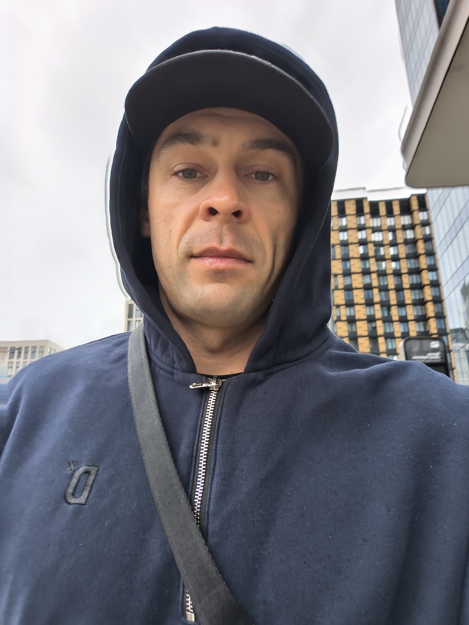 A man in a dark blue hooded sweatshirt and a black baseball cap is standing in front of a building in Moscow, Russia. The sweatshirt has a zipper down the front and a black strap is visible across his chest. The building behind him is modern, with a grid-like pattern of windows and dark orange panels. The sky is overcast.