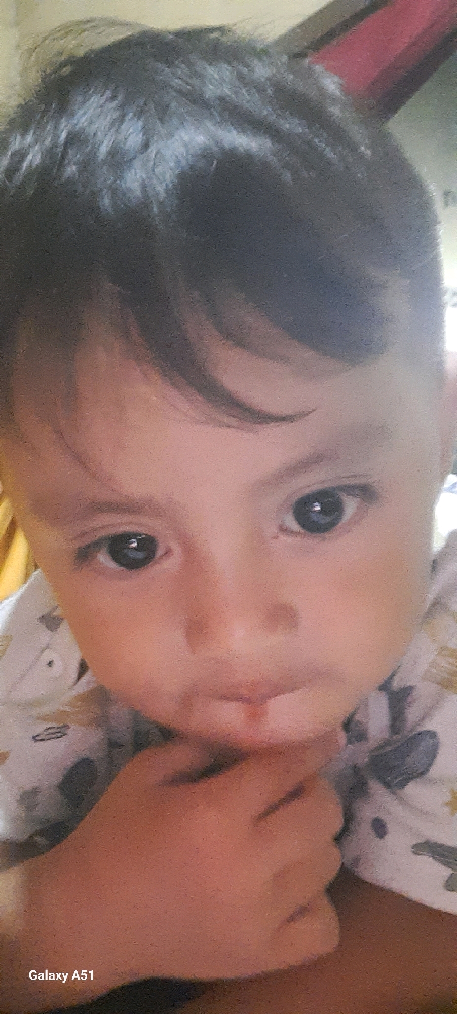 The image is a close-up portrait of a young child's face, taken indoors. The child has dark hair, fair skin, and dark eyes. Their left hand is placed near their mouth. The child is wearing a white garment with a pattern of what appears to be cartoon characters or drawings. The background is blurred, suggesting an indoor setting with some objects that are not clearly discernible. A portion of the bottom left corner of the image shows the text "Galaxy A51", indicating the device used for capture. There is no visible information or context in the image that would allow for the identification of the location within the city of Ploso, Indonesia.