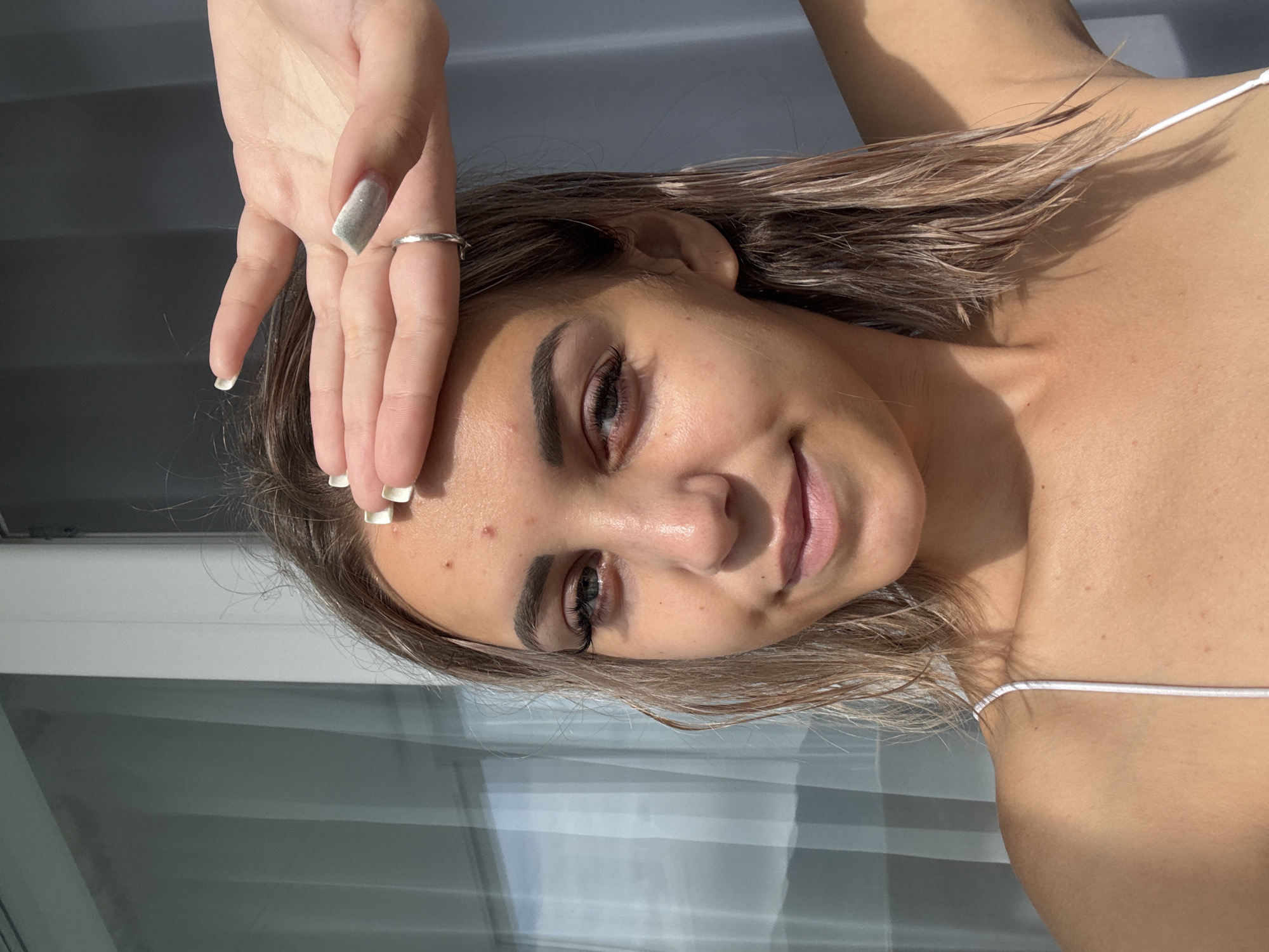 A young woman with shoulder-length blonde hair, wearing a white spaghetti strap top, is shown from the chest up. She is holding her left hand over her forehead, obscuring her eyes. She has on light makeup and a silver ring is visible on her left ring finger. Several pimples are present on her forehead. The background consists of a window with vertical blinds. There is no visible information about the location in the city of Kholmsk, Russia.