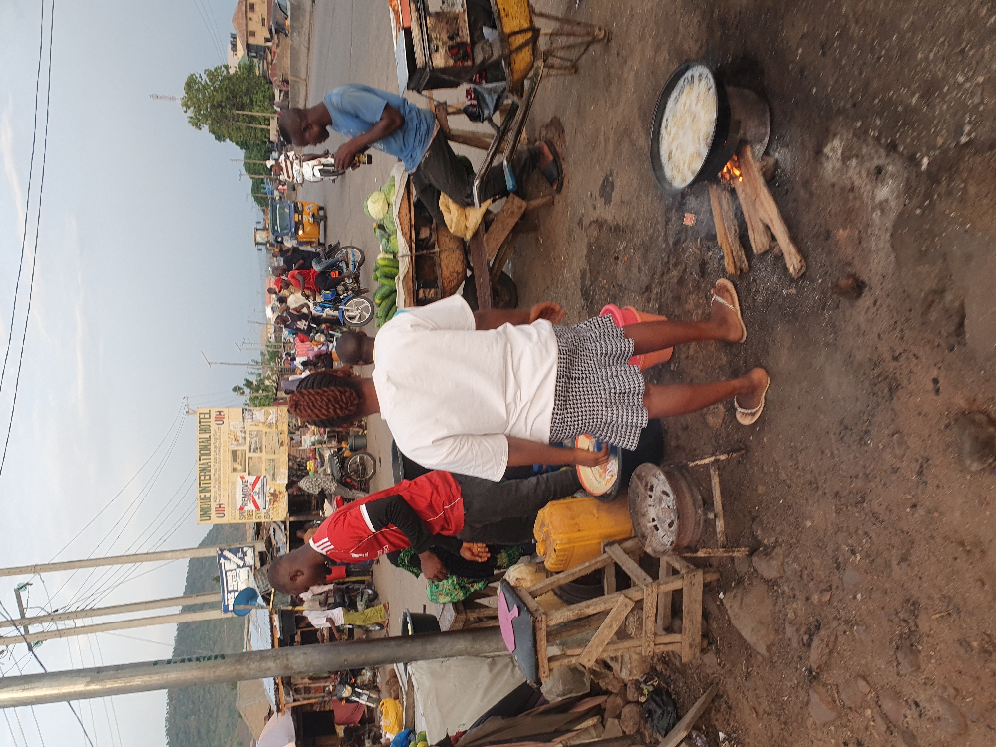 The image captures an outdoor scene along a street in Lokoja, Nigeria. A person is cooking a large pot of food over a wood fire on the bare ground. Two people, one in a white shirt and the other in a red shirt, stand nearby with various containers and objects around them, including what appears to be a wooden crate and a yellow plastic container. In the background, another person in a blue shirt is sitting on a vegetable stand beside a road with traffic and motorcycles. The "UNIQUE INTERNATIONAL HOTEL" sign is visible across the road, along with the hills in the distance. The overall scene conveys a busy, makeshift roadside cooking and trading environment.