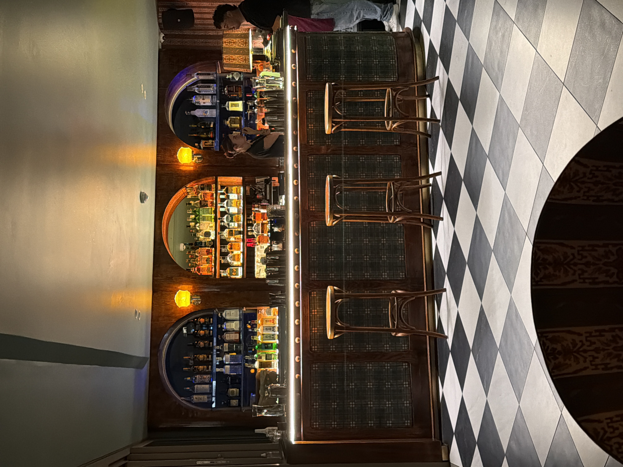This image captures an interior view of a stylish bar or lounge, likely in Cape Town, South Africa, illuminated for an evening or nighttime setting. The focal point is a long, dark-wood bar counter, featuring an integrated strip of bright white LED lighting along its top edge and textured, possibly padded, front panels. Three wooden bar stools with round seats are arranged in front of the counter, inviting patrons to sit.
Behind the bar, two individuals, presumably bartenders, are at work. A male figure is visible towards the far end of the bar, while a female bartender is positioned closer, appearing to observe something on the counter.
The back wall of the bar is adorned with three large, arched alcoves. Each alcove is fitted with multiple shelves generously stocked with an array of spirit and liquor bottles, brightly illuminated from within to showcase the collection. Two small, yellow-glowing wall sconces are installed between the lower and middle alcoves, contributing to the warm ambient light. A dark speaker is mounted on a higher shelf in the background.
The floor to the right of the bar displays a striking black and white checkered pattern, laid out diagonally. A portion of a round, dark table with an intricately patterned border is visible in the bottom right corner of the frame. No discernible text is clearly legible on any of the bottles or other elements within the scene. The overall ambiance suggests a sophisticated and inviting establishment.