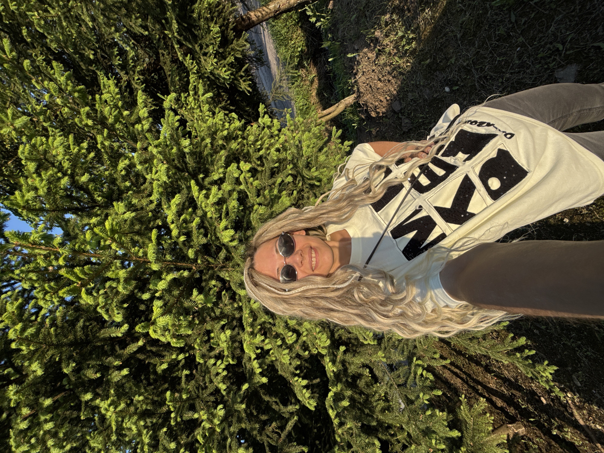 A woman with long blonde hair wearing sunglasses and a light-colored jacket with large black lettering is lying on the ground near a large evergreen tree, possibly a spruce or fir. The environment is grassy, suggesting an outdoor location, possibly a park or wooded area in Rusanovo, Russia. A paved area is visible in the background. The photo is taken from a high angle, looking down on the subject.
