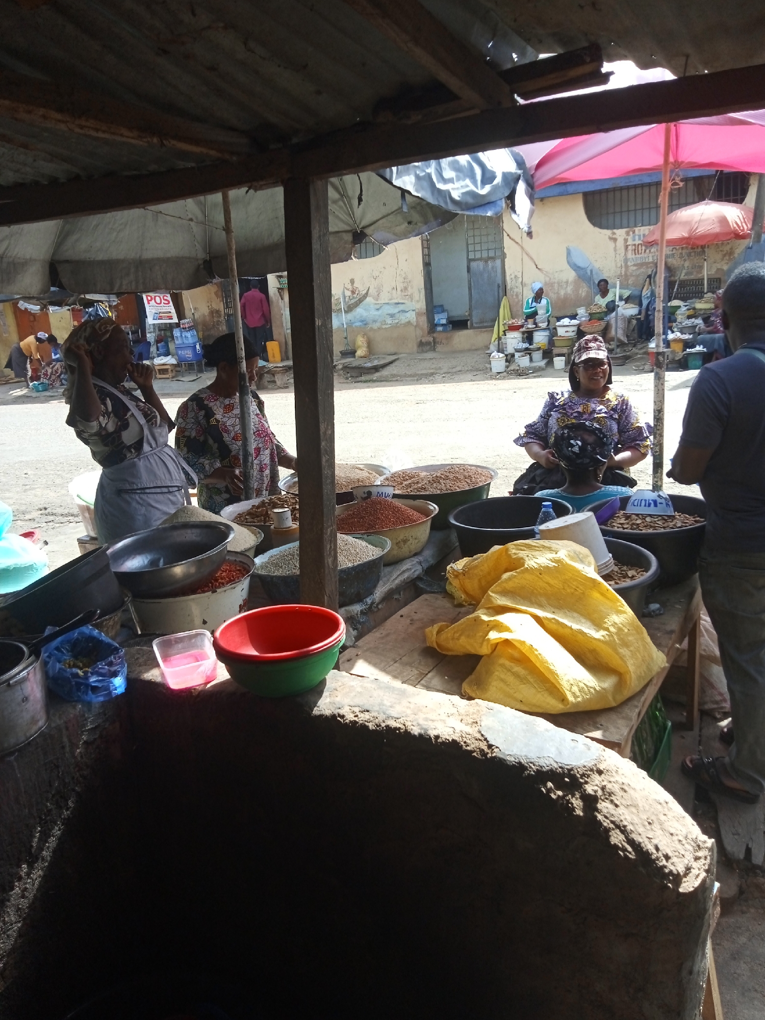 Here is a detailed description of the image:
**Location and Environment:** The image is taken at an outdoor market in an unspecified city. The market appears to be bustling with activity. The location is possibly near a street with buildings in the background, some shaded by makeshift shelters or umbrellas.
**People:**
- Several individuals are visible at the market.
- One woman is standing at a stall filled with goods.
- Another woman is attending to a child, while a man appears to be interacting with her.
- Other figures are discernible in the background, including vendors and customers.
**Subjects and Objects:**
- The main focus is a stall laden with goods.
- Various types of grains, seeds, or nuts are displayed in multiple containers.
- Cooking utensils like bowls and pots are scattered around the stall.
- A yellow bag is placed on the counter.
**Actions and Interactions:**
- Vendors are likely selling their goods to customers.
- Interactions between vendors and customers are likely occurring.
**Overall Impression:** The scene depicts a typical day at an outdoor market, showcasing the vibrant commercial activity and everyday life within the community.