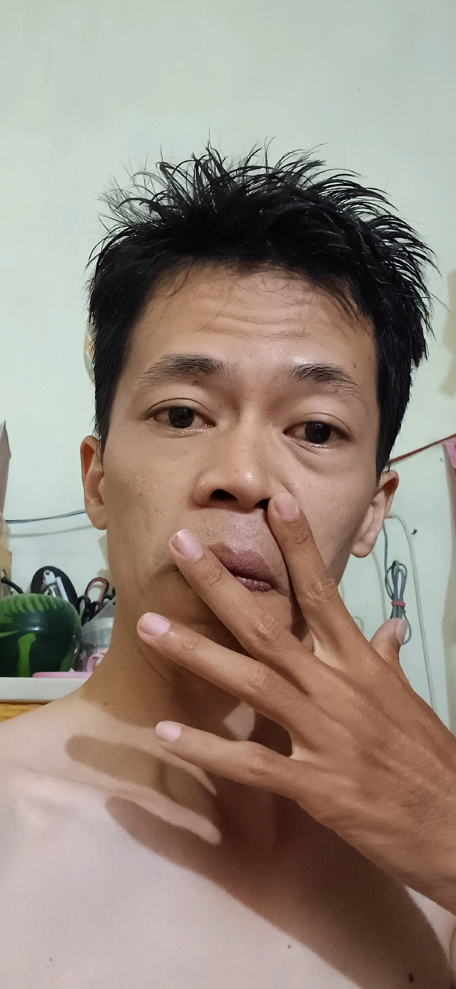 A close-up, head-and-shoulders selfie shows a person's face, with their hand covering their mouth and nose. The person has dark, spiky hair, brown eyes, and light skin. The background is a plain, light-colored wall with some wires and objects visible on shelves to the left and right. The lighting suggests an indoor setting. The image appears to have been taken in Wonokerso, Indonesia.
