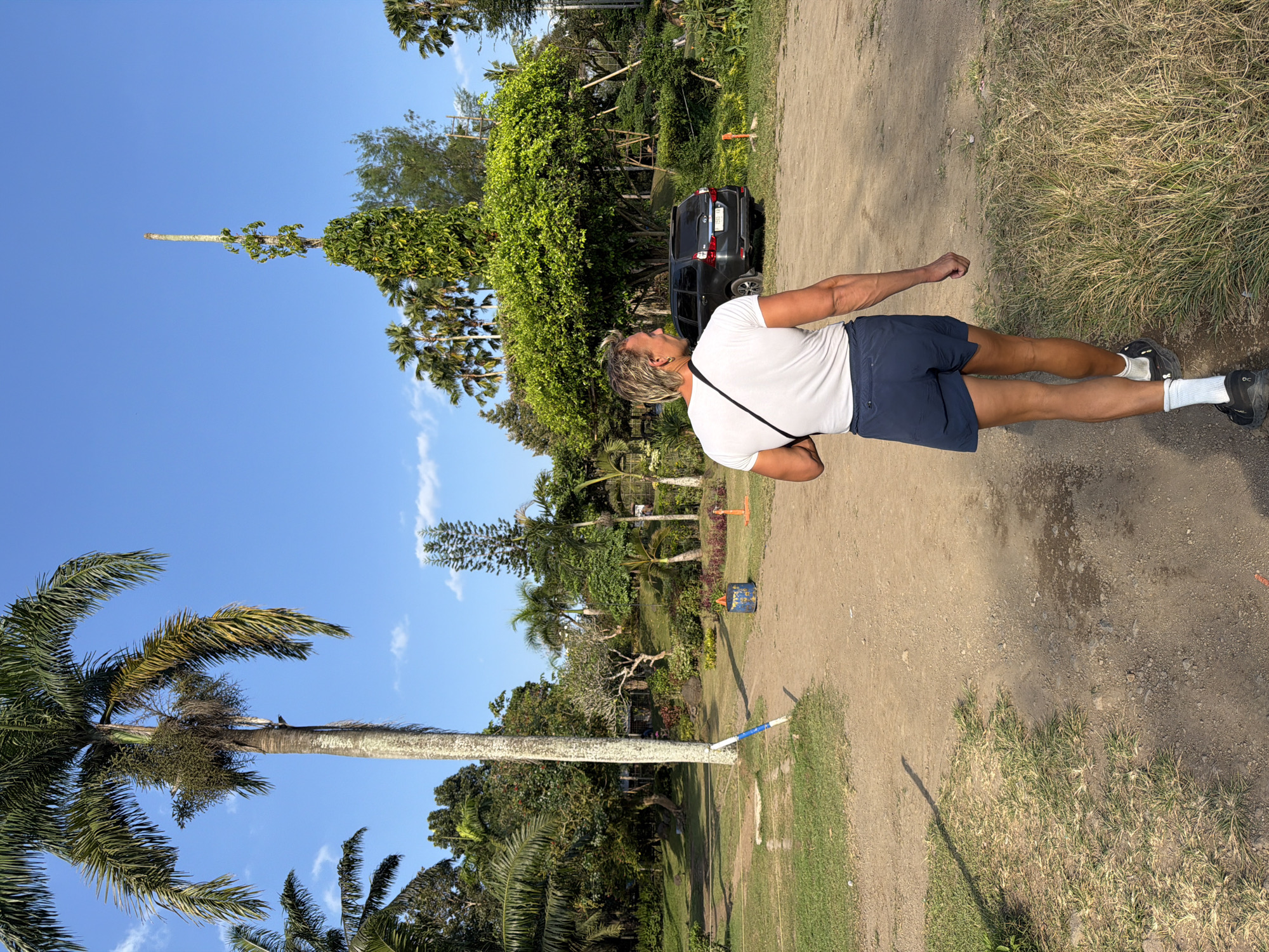 A man with blonde hair and muscular arms walks away from the camera on a dirt path. He is wearing a white t-shirt, navy blue shorts, white socks, and black athletic shoes. A black bag with a strap is slung across his chest. The path is surrounded by grass and dirt. In the background, there are numerous palm trees and other lush green vegetation under a clear blue sky with a few scattered clouds. A dark-colored SUV is parked on the right side of the path. A tall, slender pole with green vines wrapped around it is visible in the distance.