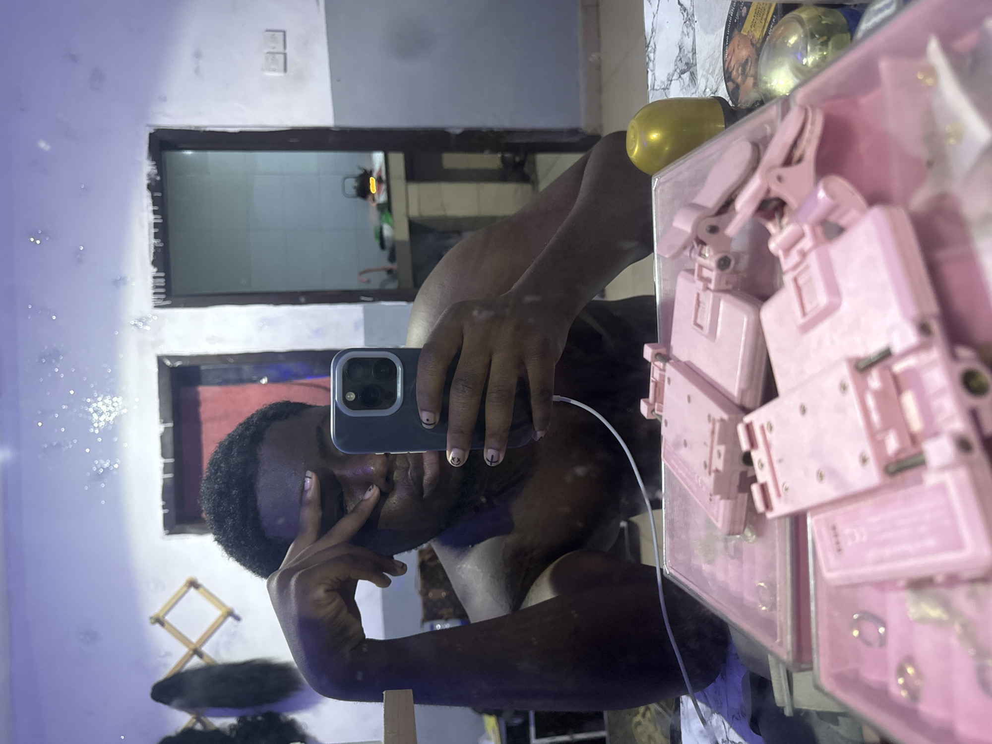 A person with dark hair and skin is taking a selfie in a bathroom. The person's face is partially obscured by their hand holding a smartphone. The phone case is dark. The person has small designs on their fingernails. A white charging cable connects to the phone. In the reflection, an open doorway is visible. A device is plugged into the wall behind the doorway, possibly a small generator. A pink container filled with what appear to be pink batteries or battery-like objects is on the right side of the frame, slightly obstructing the view. An unidentified gold spherical object is partially visible above the pink container.
The environment is a tiled bathroom. The wall reflects sparkling particles and lighting, obscuring details. A collapsible wooden rack or dryer is visible. The location is Ipetumodu, Nigeria, based on the available information.
