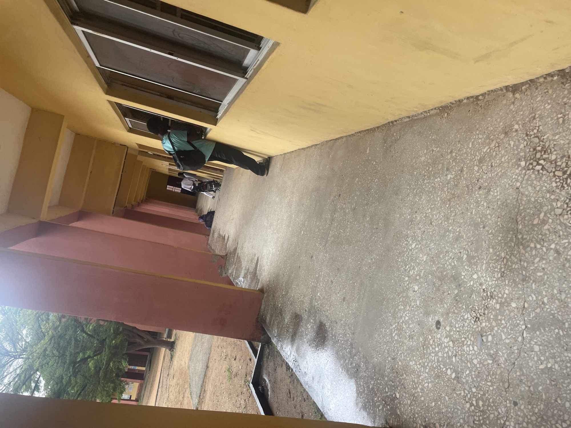 The image shows a student looking out of a window in Lokoja, Nigeria. The student is wearing a teal shirt and black pants, and has a black backpack. They are leaning out of a window of a yellow building with red accents. The ground in front of the building is a wet, grey concrete walkway. In the background, there are trees and other buildings. There are a few other people in the background, one of whom is on a motorcycle.