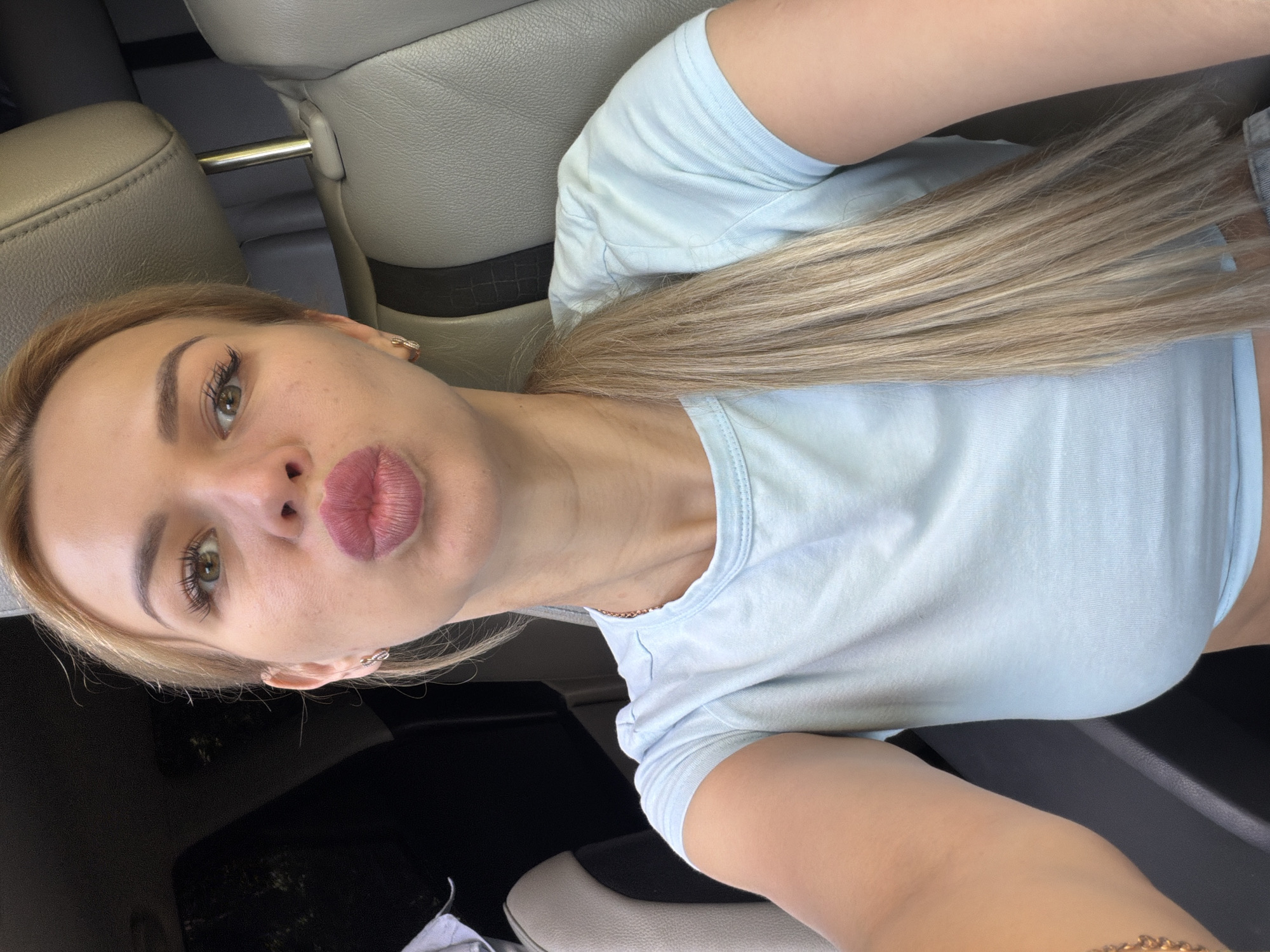 A blonde woman is sitting in the driver's seat of a car, likely in Kyiv, Ukraine. She is looking at the camera and puckering her lips for a selfie. She is wearing a light blue t-shirt and her long blonde hair is pulled back in a ponytail. The interior of the car is visible behind her, with the headrest and seats of a car.