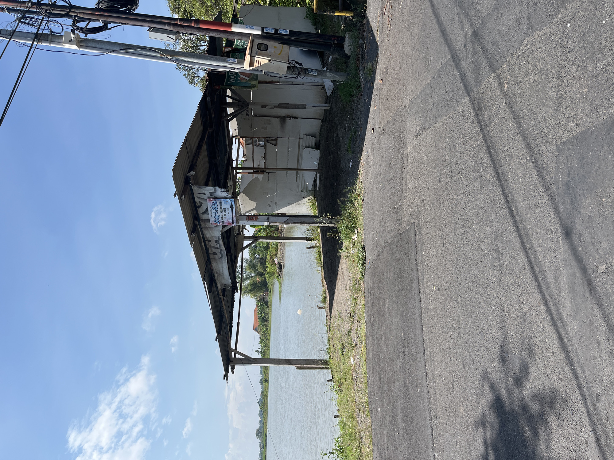 The image shows a rustic, open-sided structure with a corrugated metal roof, situated next to a body of water. The structure appears to be made of wood and concrete and shows signs of disrepair. There is a sign hanging from the roof with text on it. The surrounding environment includes grass, dirt, a road in the foreground, and trees and houses in the background. The sky is blue with scattered clouds. Utility poles with wires are visible to the right of the structure. The location is identified as Pesawahan, Indonesia.