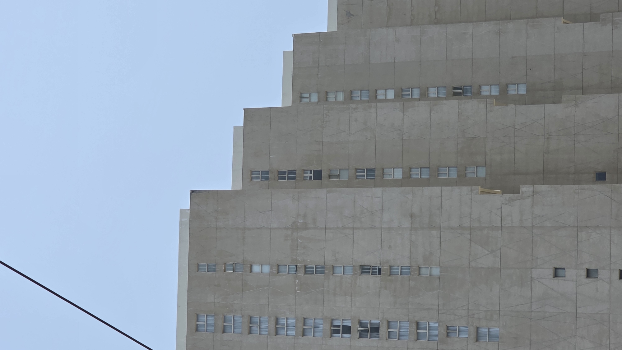 A partial view of a stepped concrete building against a clear blue sky is shown. The building appears to be modern architecture with a stacked design. The facade is characterized by repetitive square windows arranged in rows across each level. A black wire or cable extends diagonally across the lower left corner of the image, obscuring a portion of the sky. The building is the Khanh Hoa Provincial General Hospital in Nha Trang, Vietnam.
