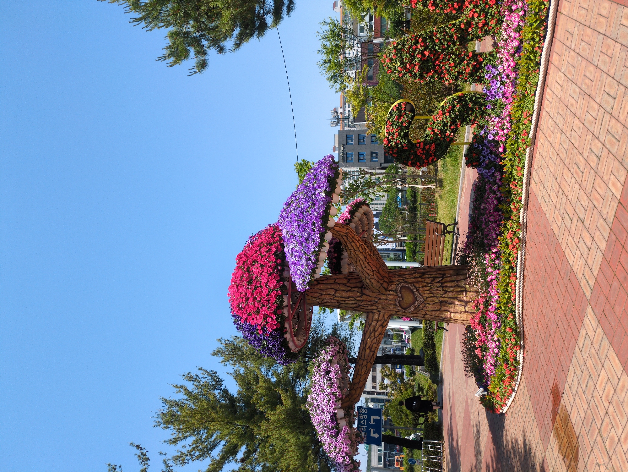 The image shows a park in Daejeon, South Korea. The focus is on a decorative tree-like structure adorned with colorful flowers. The "tree" has a brown "trunk" and "branches" that hold clusters of red, pink, and purple flowers. The trunk features a heart-shaped design. Surrounding the base of the tree is a garden bed filled with more flowers.
Behind the tree, there's a park setting with a bench and additional floral displays. In the background, there are buildings, typical of an urban area. A paved pathway leads through the park. A person can be seen walking along the path. The sky is clear and blue. A street sign is visible in the lower portion of the image. The overall scene suggests a well-maintained park area within the city.