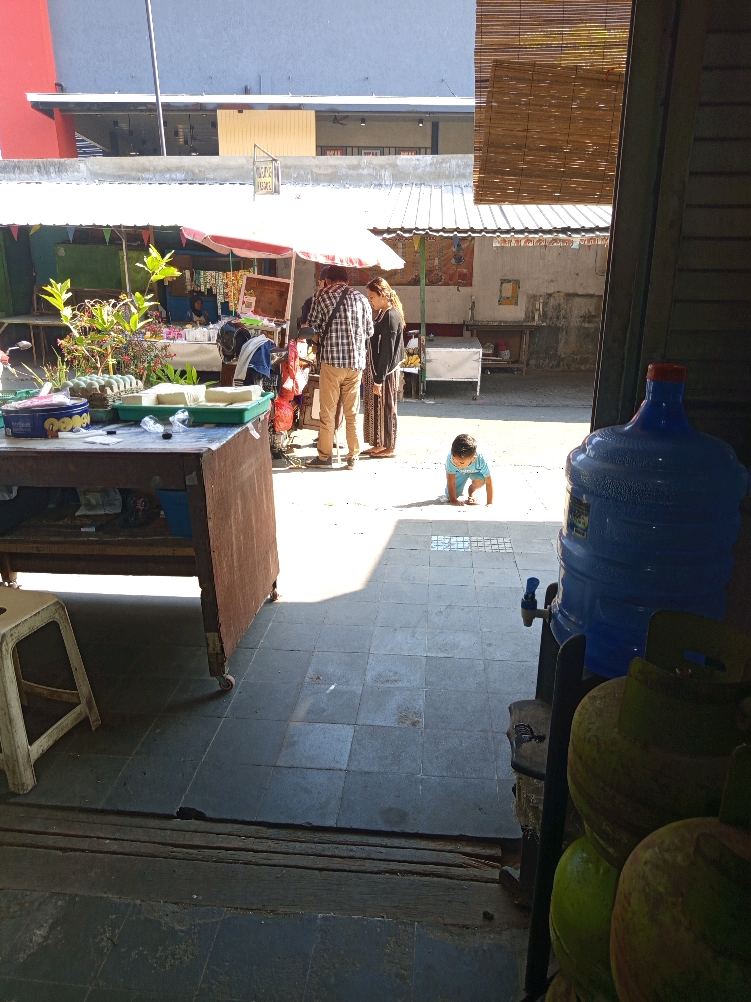 In a sunlit outdoor market stall area, possibly in Malang, Indonesia, a young child in a light blue shirt is crawling on a tiled floor. The child is positioned on the left side of a large blue water dispenser with a red cap and a blue spigot. To the right of the child and dispenser are several large, round objects that appear to be propane gas tanks. In the background, a table laden with produce, including tofu and eggs, is set up under a white awning. Two adults, one in a plaid shirt and khaki pants and the other in a dark outfit, are standing near the table, seemingly engaged in commerce. A motorcycle is parked behind them. The scene suggests a busy marketplace with goods for sale and everyday life unfolding. The architecture in the background indicates a multi-story building.