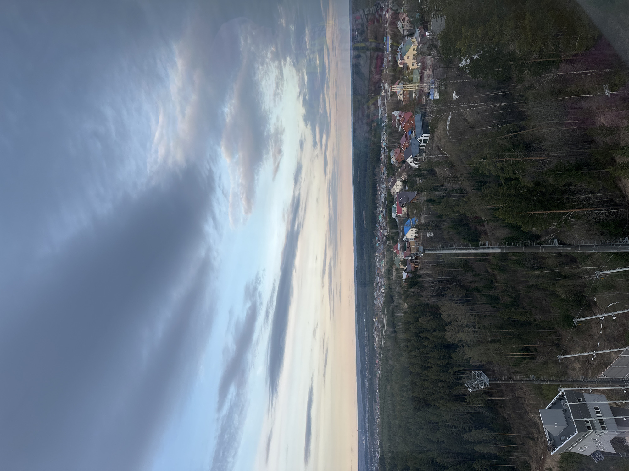 The image shows an elevated view of Прикамский, Russia. The right side of the frame displays a residential area with houses of varying colors and sizes nestled along the edge of a dense forest. Wires and utility poles are visible amidst the trees. Adjacent to the houses, a long, elevated walkway or bridge-like structure extends into the forest. The lower right corner features a modern building with a grey roof and multiple windows. The left side of the image is dominated by a cloudy sky with shades of grey, white, and hints of blue and peach near the horizon. The sky suggests either sunrise or sunset conditions. The division between the sky and the landscape appears slightly blurred, possibly due to the angle of the photograph or atmospheric conditions.