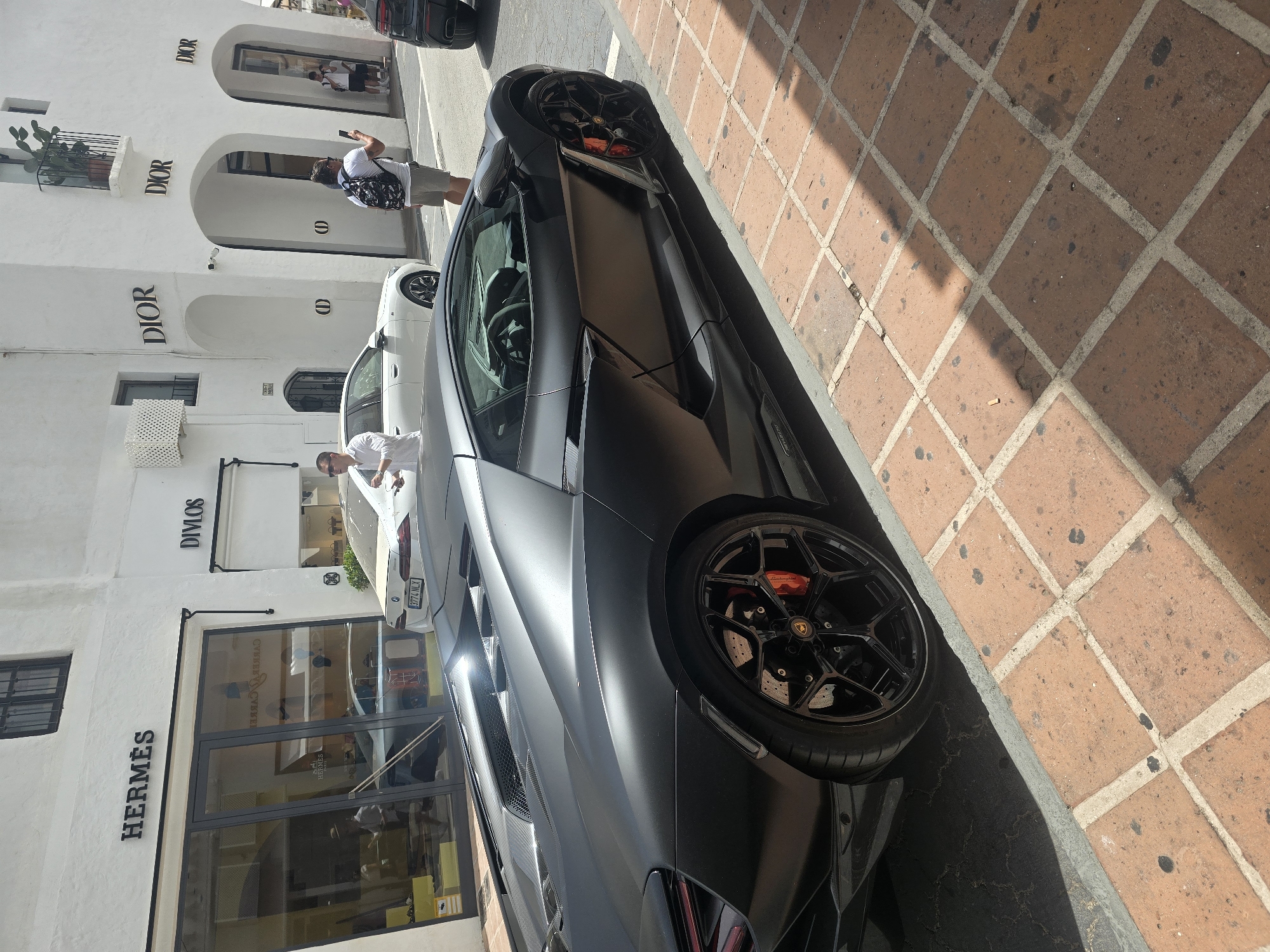In Marbella, Spain, a matte black Lamborghini is parked on a cobblestone sidewalk adjacent to a street. The car's distinctive features, including its low profile, aggressive styling, and bright red brake calipers, are prominent. Behind the car, a white building with the logos of luxury brands such as "HERMES" and "DIOR" is visible, suggesting a high-end shopping district. Several people are present, including a man taking a picture with his phone and another individual standing near a white car. The overall scene captures a moment of luxury and urban life in a Mediterranean setting.