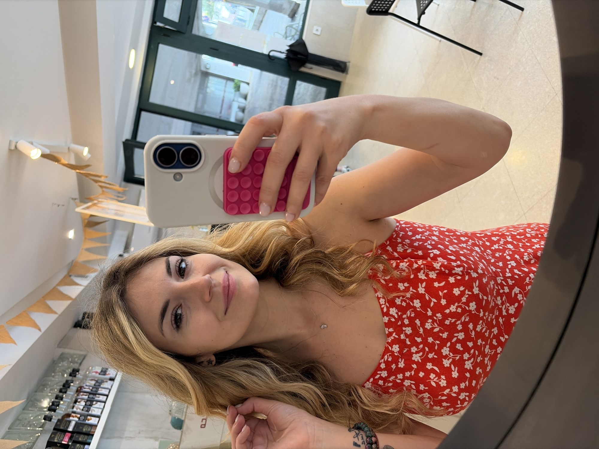 A young woman is taking a selfie in what appears to be a beauty salon or shop in Budva, Montenegro. She is wearing a red floral print dress and holding a white smartphone with a pink pop socket. Her blond hair is wavy and falls around her shoulders. She has a tattoo on her left wrist and is wearing a necklace. Behind her is a window overlooking a street scene, and to the left, there are shelves displaying various products. Above the shelves, there is a string of triangular pennants. The image is taken in a mirror, so the person and scene are reflected. A chair can be seen in the background.