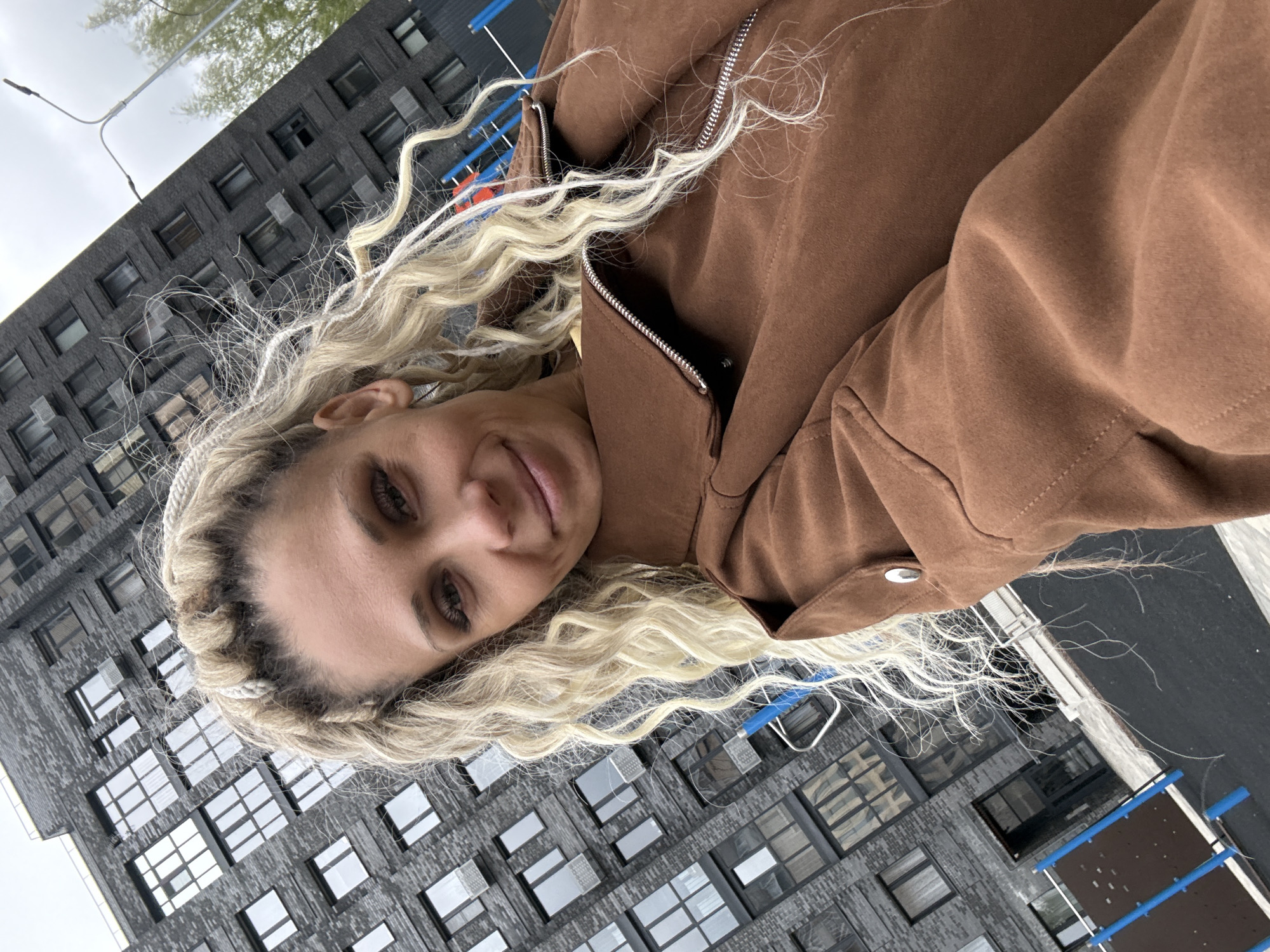 A woman with blonde braided hair, wearing a brown jacket with a partially zipped silver zipper, is outdoors. The background shows a dark grey building with rows of windows, and a piece of playground equipment with blue bars is visible on the right. The image appears to be taken in Rusanovo, Russia.