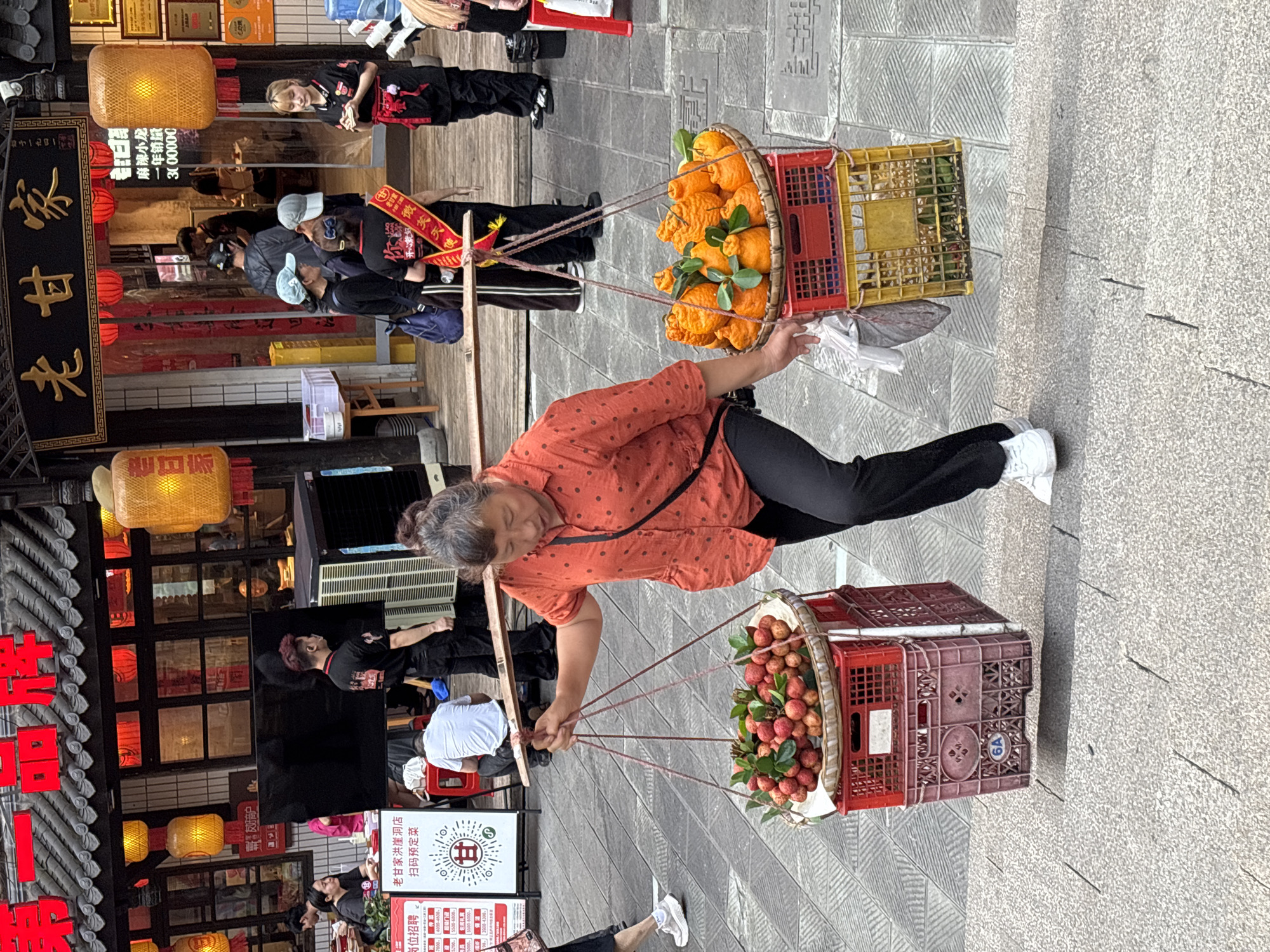 The image shows an older woman in the Yuzhong District, China, carrying fruit on a shoulder pole. She wears a coral-colored, polka-dot shirt, black pants, and white sneakers. Two baskets, each containing different types of fruit, are attached to the pole. One basket holds oranges with green leaves, while the other contains smaller red fruits, possibly lychees or longans. The baskets are placed on top of stacked plastic crates. She is on a stone-paved street alongside a building with traditional Chinese architecture. The building has red lettering on the facade, and lanterns hang outside. People, including a group with red sashes, walk by. A sign near the woman has Chinese characters, including job listings.