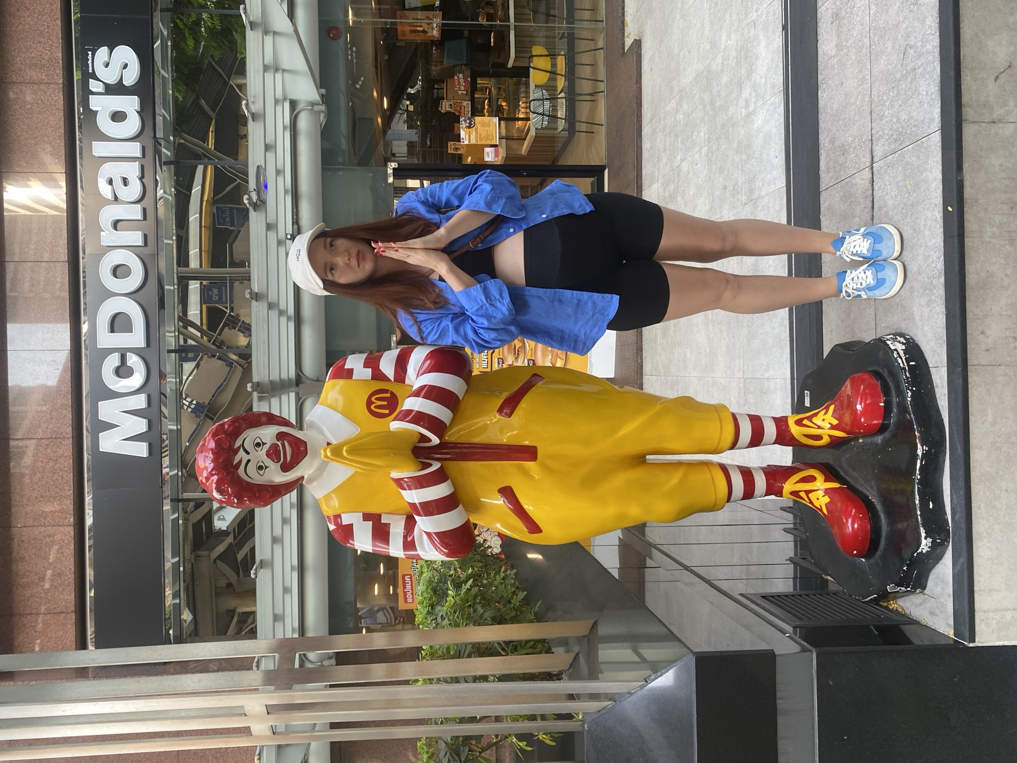 A young woman and a Ronald McDonald statue are depicted side-by-side in a brightly lit outdoor setting, presumably during the daytime. The woman has long brown hair and is wearing a white baseball cap, a blue button-up shirt over a black crop top, black shorts, and light blue sneakers. She stands with her hands pressed together in a traditional Thai "wai" greeting, looking towards the camera with a neutral expression.
Beside her, the Ronald McDonald statue, in its classic yellow jumpsuit with red and white striped sleeves and red shoes, also performs a "wai" gesture. The statue features its characteristic red hair and clown makeup. A golden "M" logo for McDonald's is visible on its chest.
The background shows the storefront of a McDonald's restaurant. The prominent "McDonald's" logo is displayed on the building. Through the glass windows, parts of the restaurant's interior are visible. Several signs with Thai text are present, including "แอปแมค" (App Mac) and "1 พอยต์" (1 point), indicating loyalty program information. The presence of these Thai language cues and the culturally specific "wai" gesture by Ronald McDonald confirm the location to be Bangkok, Thailand. The ground is paved with light-colored tiles, and some foliage is visible near the base of the statue.