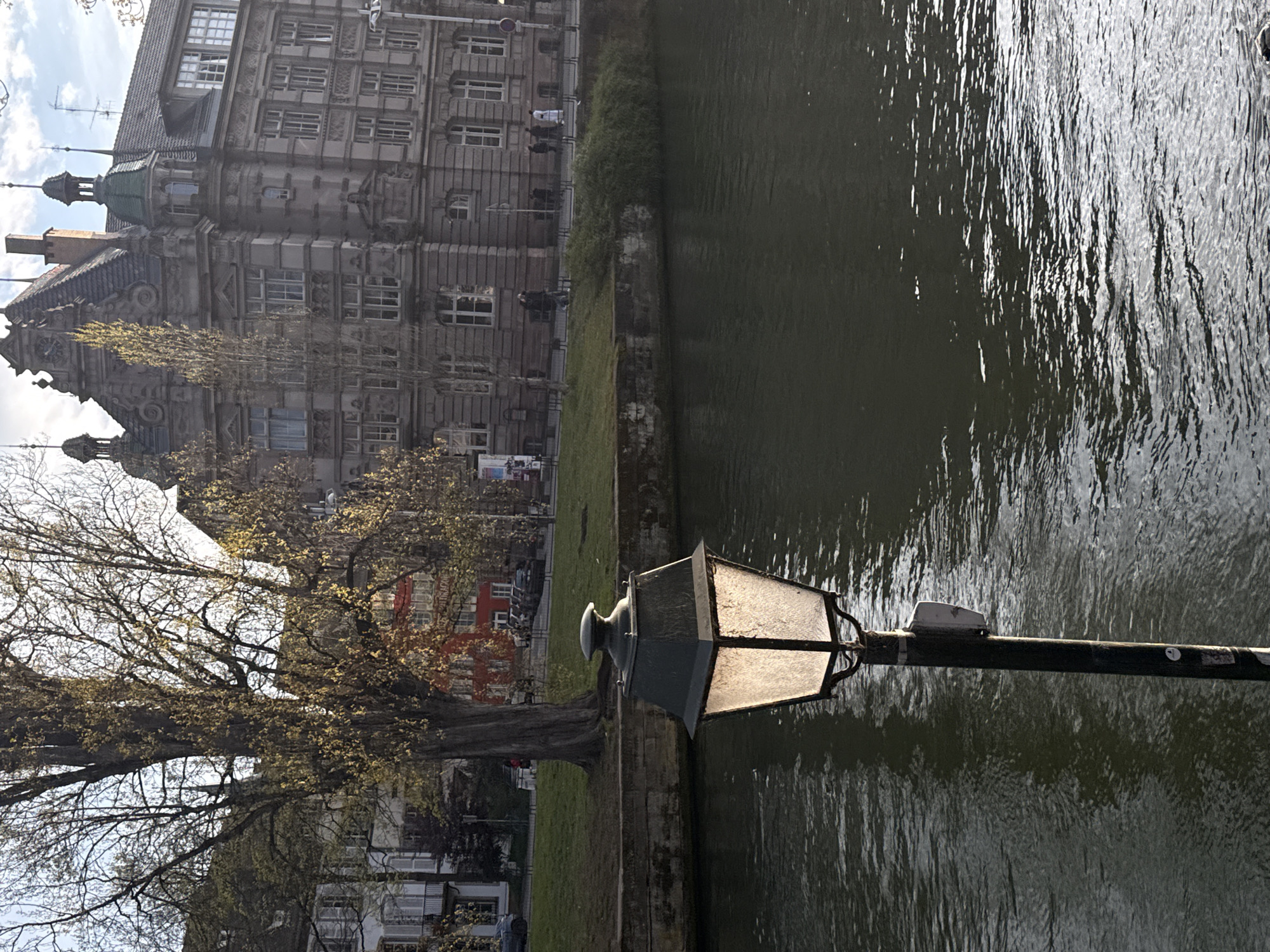 The image depicts a waterway in Strasbourg, France, with a prominent, illuminated lamppost in the foreground extending horizontally into the scene. To the left of the lamppost is a grassy bank with a bare tree. Behind the tree, buildings are visible, including an elaborate multi-story building with decorative architectural features. People are walking along the path in front of this building. The waterway occupies the right side of the frame, with reflections visible on the water's surface. The sky is partially visible, and the lighting suggests it is daytime. The buildings' architecture is characteristic of Strasbourg.