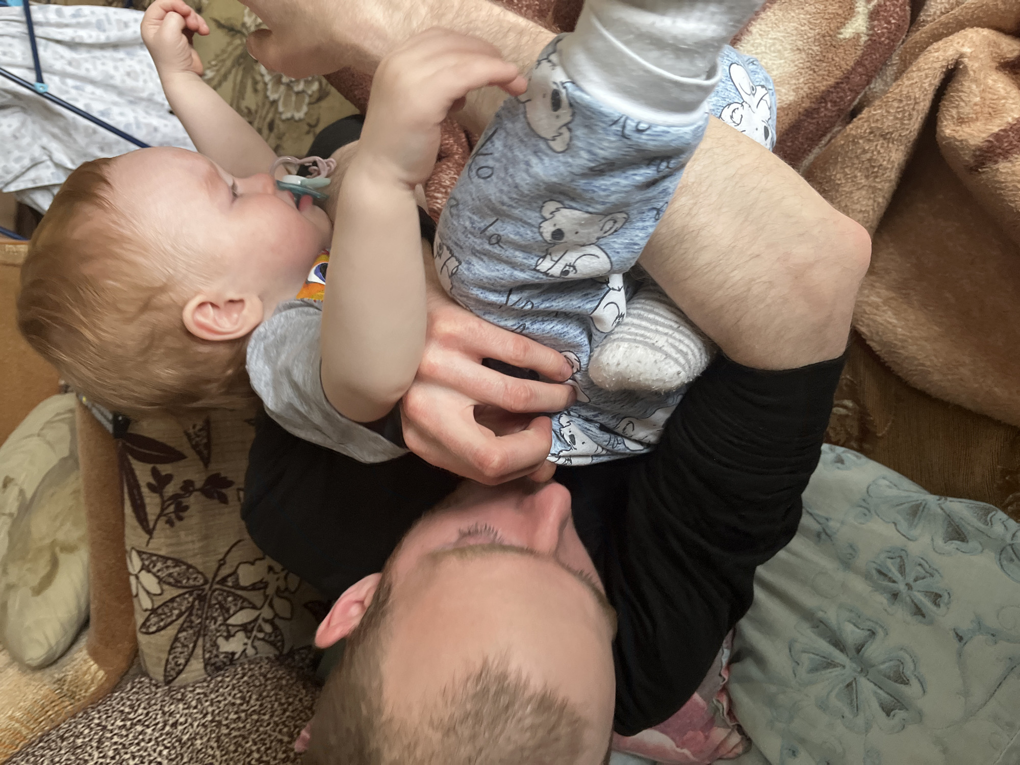 A man is lying down with a baby held up above him. The baby is wearing a gray shirt and blue pajamas with koala bears printed on them, and is holding their legs up in the air. The baby has a pacifier in their mouth. The man has short hair and is wearing a black shirt. The man is either sitting or lying on a patterned couch with an afghan. The picture was taken in an indoor setting. It is not possible to ascertain the location in Сосново, Russia, based on the content of this image.