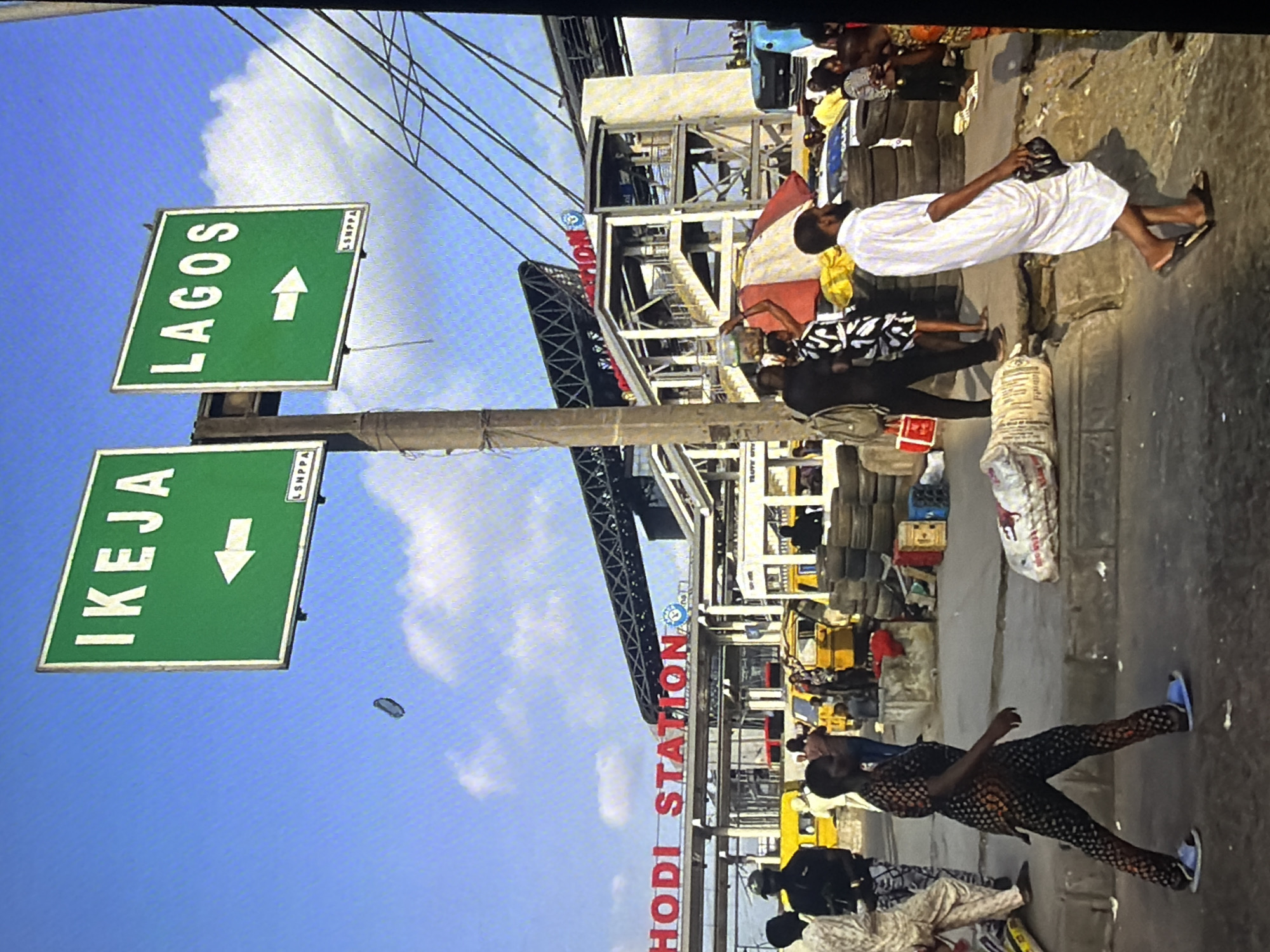 The image depicts a street scene in Bukuru, Nigeria, under a bright, partly cloudy sky. Two green directional signs, one indicating "IKEJA" with a left arrow and the other "LAGOS" with a right arrow, are prominently displayed above. In the background, a multi-level structure with an elevated walkway or bridge is visible, identified by red lettering that reads "RHODI STATION". Below the signs and the station, a busy street scene unfolds. Yellow minibuses, commonly known as "danfos," are parked and in motion, suggesting a public transport hub. A variety of people are present, some walking, some standing. One person in the foreground wears a patterned outfit and blue sandals, walking from left to right. Another individual, dressed in a white robe and black sandals, is also walking rightward, carrying a dark bag. Piles of used tires are stacked along the roadside, and various goods, including sacks and crates, are scattered around. The overall impression is of a bustling urban environment.