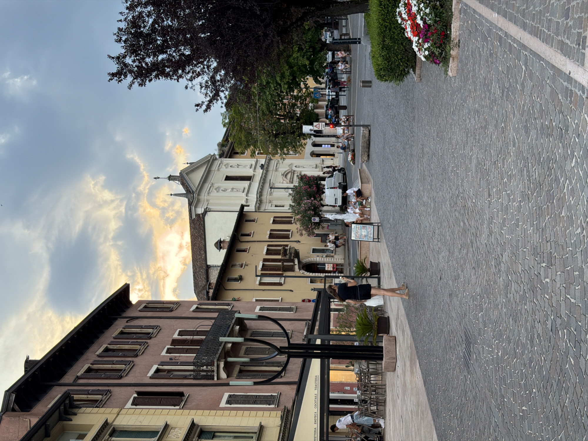 The image shows a street scene in the city of Salò, Italy. The foreground is dominated by a cobblestone pavement. To the left, a pink building with closed brown shutters lines the street, with an awning featuring the text "CAFFETTERIA ENOTECA COCKTAILS TOASTERIA". An ornate, dark metal lamppost stands on the sidewalk. Further down the street, a yellow building with similar shutters is visible, and beyond that, a white church with a prominent facade and a cross atop its roof. People are scattered throughout the scene: a woman in a black dress is walking towards the right, a man in a white t-shirt is standing by tables and chairs outside the cafe, and several individuals are seated near a fountain in the mid-ground. Cars are parked along the street in the background. A dense, dark-leaved tree is on the right side of the image, and a flowerbed with red and white flowers is at the bottom right. The sky is overcast with patches of sunlight breaking through, suggesting it is late afternoon or early evening.