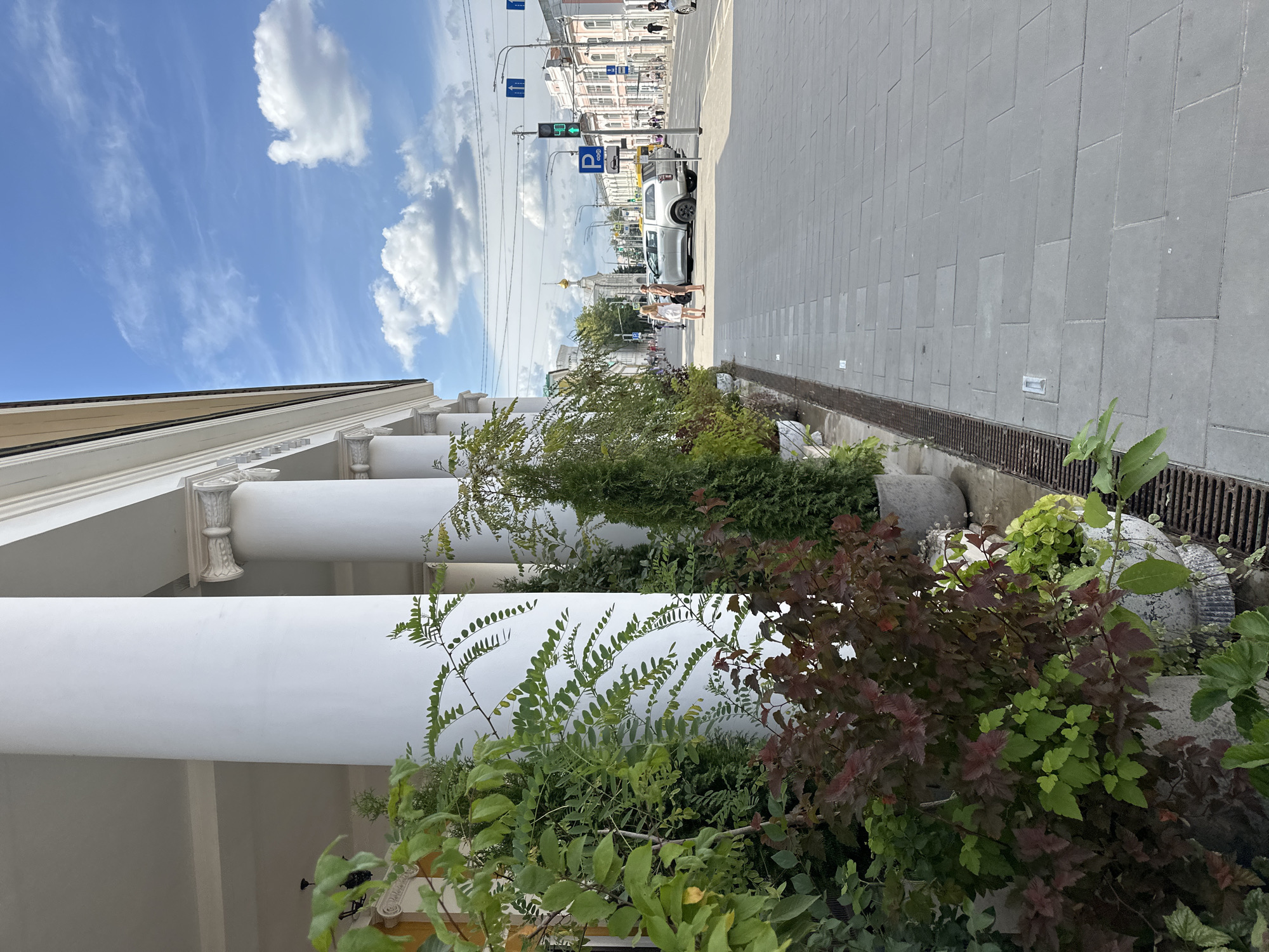 The image depicts a street scene in Golovinskoe, Russia, characterized by classical architecture and a sunny day. On the left, a colonnade of white columns lines the building, with potted plants, including green foliage and dark red leaves, placed in front of them. A drain grate is visible along the edge of the sidewalk.
The street itself is paved with grey rectangular tiles. A silver SUV is parked on the right side of the street, facing away from the camera. Two individuals, a man and a woman, are standing near the SUV, appearing to converse.
Further down the street, traffic lights are visible, along with road signs, including a blue "P" indicating parking. Buildings with light-colored facades line the street on the right. In the distance, a church with a golden dome stands prominently, suggesting a historical or religious site. Power lines and streetlights are also present. The sky is blue with scattered white clouds, indicating pleasant weather. The overall scene captures a moment of urban life with a blend of historical and modern elements.