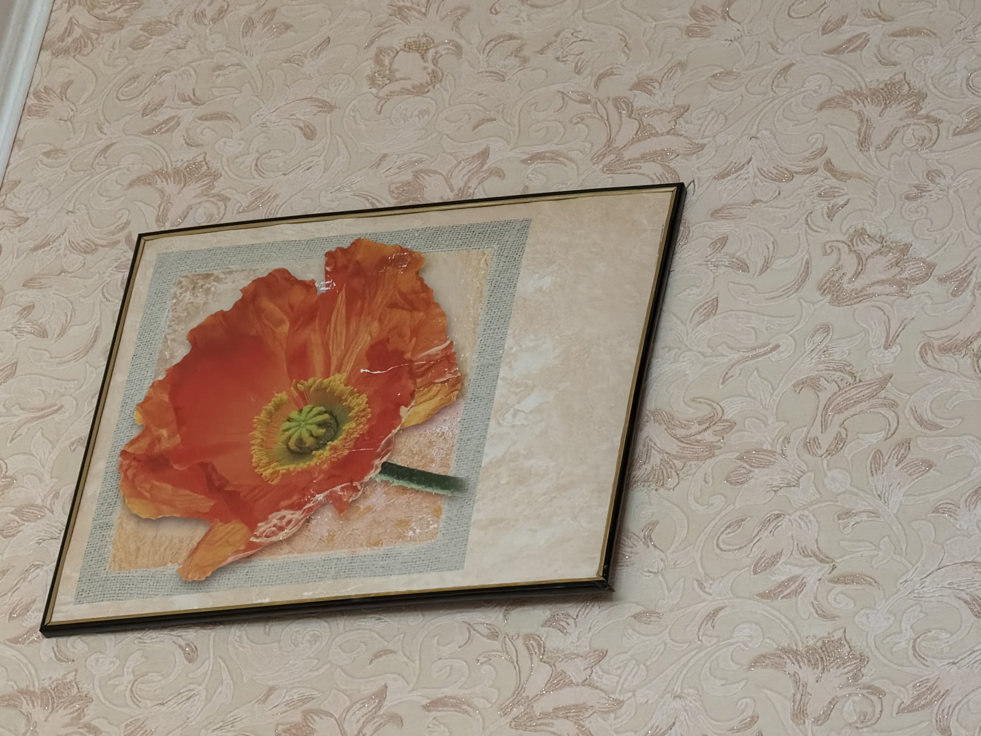 A framed picture of an orange poppy hangs on a wall with floral wallpaper. The picture is square with a thin black frame. The poppy fills most of the picture, with its petals a vibrant orange and its center a yellow-green. The background of the picture appears to be a textured surface with hints of cream and grey. The wallpaper is a pale cream color with a raised floral pattern in a slightly darker cream and some subtle pink accents. The overall impression is of a domestic interior.