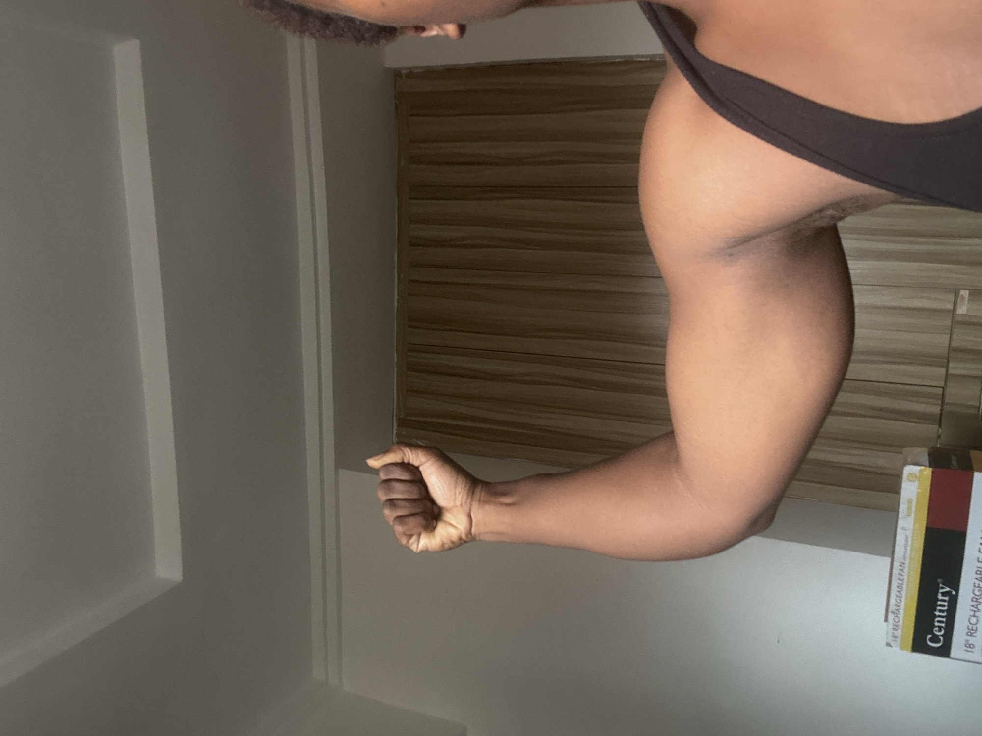 The image, rotated 90 degrees counter-clockwise, presents a close-up of a person's upper body, specifically their arm and side. The subject, who has a dark skin tone, is flexing their right bicep, with their hand forming a tight fist. They are wearing a dark-colored, possibly black, tank top or undergarment, which partially reveals their torso and shoulder. Some body hair is visible in the armpit area.
The background features an indoor setting. To the left, a plain, light-colored wall is visible, adorned with subtle vertical rectangular molding or paneling. To the right, behind the person's upper arm, there is a wall or piece of furniture with horizontal wooden slats, providing a warm, textured contrast.
At the bottom right corner of the frame, a stack of items resembling product boxes or large books is visible. Clear text on one of these items reads "18" RECHARGEABLE FAN" and the brand "Century." Another instance of "18" RECHARGEABLE FAN" is also partially visible. The lighting appears to be ambient indoor light, casting soft shadows. The overall scene suggests a personal moment, perhaps a display of physical fitness or strength, taking place in an indoor environment in Abuja, Nigeria.