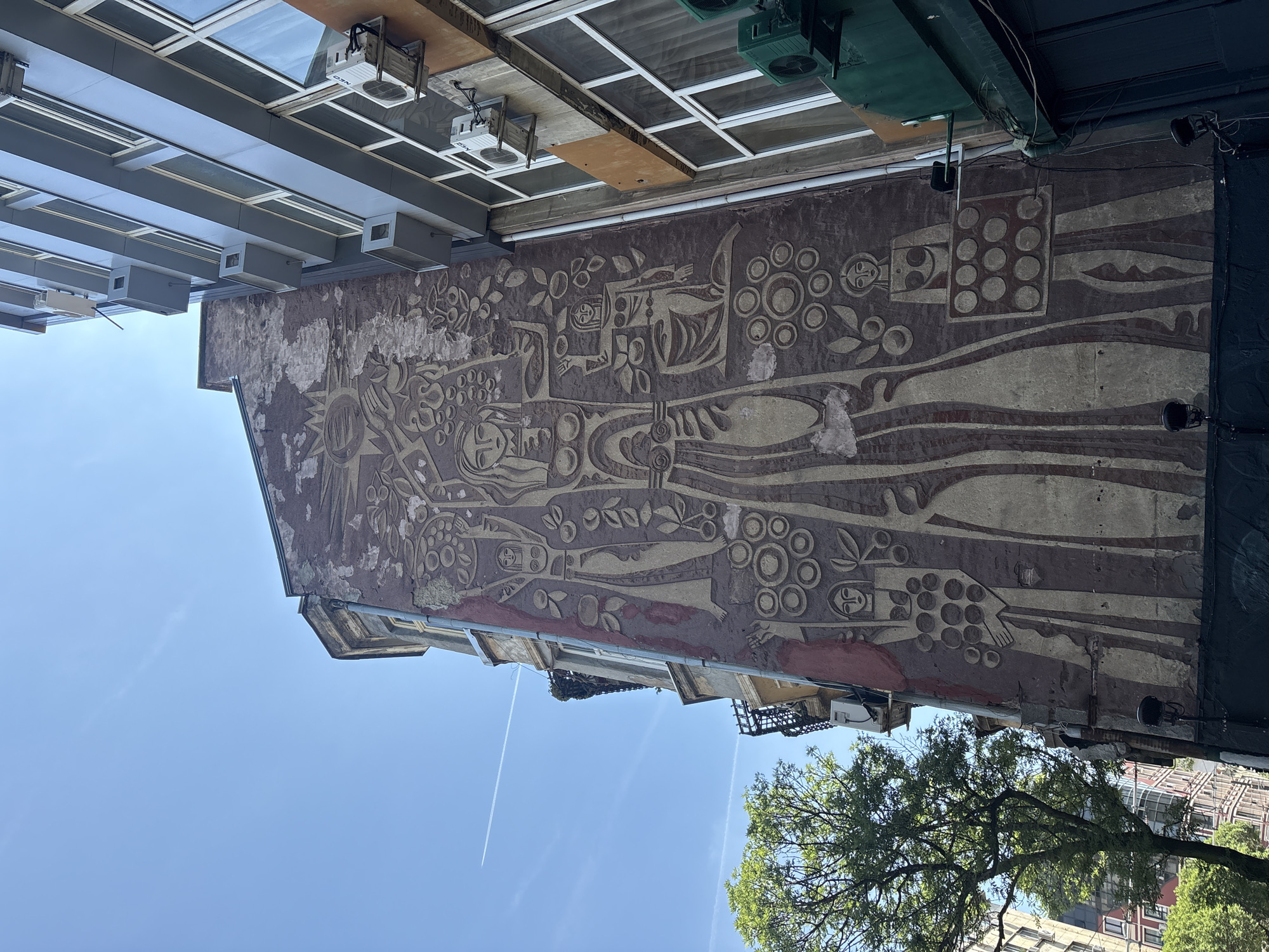 A large, weathered bas-relief or mural dominates the side of an old building in Sofia, Bulgaria, captured from a low angle looking upwards towards a bright blue sky. The artwork, in an abstract and stylized manner, depicts multiple human-like figures integrated with organic motifs such as leaves, branches, and clusters of circular forms resembling fruits or symbolic patterns. The figures have simplified features and appear elongated. The mural is textured, suggesting it's carved or sculpted, and shows signs of aging with sections of the surface peeling away, revealing underlying material.
Above the mural, a contrasting modern structure with a grid of glass panels, possibly a skylight or part of an upper floor, is visible. Several white rectangular units, possibly HVAC systems, are mounted beneath this structure, with "NEC" visible on some. Various pipes, cables, and dark light fixtures are also attached to the building facades. The sky is clear and bright, indicating daytime, with a distinct airplane contrail visible in the upper atmosphere. The top of a green leafy tree is seen in the lower right corner, grounding the urban scene with a natural element.