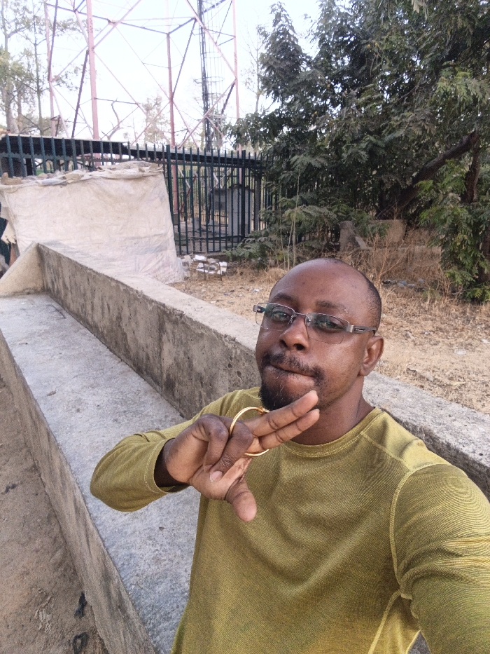 The image shows an adult male in an outdoor setting in Garun Gawa, Nigeria. The individual, with dark skin, a shaved head, and short facial hair, wears clear-framed glasses and an olive-green long-sleeved shirt. He is positioned in the lower right of the frame, facing the camera, with his right hand raised, displaying a gold-colored ring on his ring finger while forming a 'V' shape with his index and middle fingers. In the mid-ground, a light-grey concrete structure with two parallel tiers extends from the lower left. Behind this, a dark green metal fence is visible, with a light-colored tarpaulin or fabric-covered object directly behind it. The background features a large, reddish-brown metal lattice tower on the left against an overcast sky, and dense green foliage from trees and bushes on the right. The ground is dry and dusty with sparse vegetation.