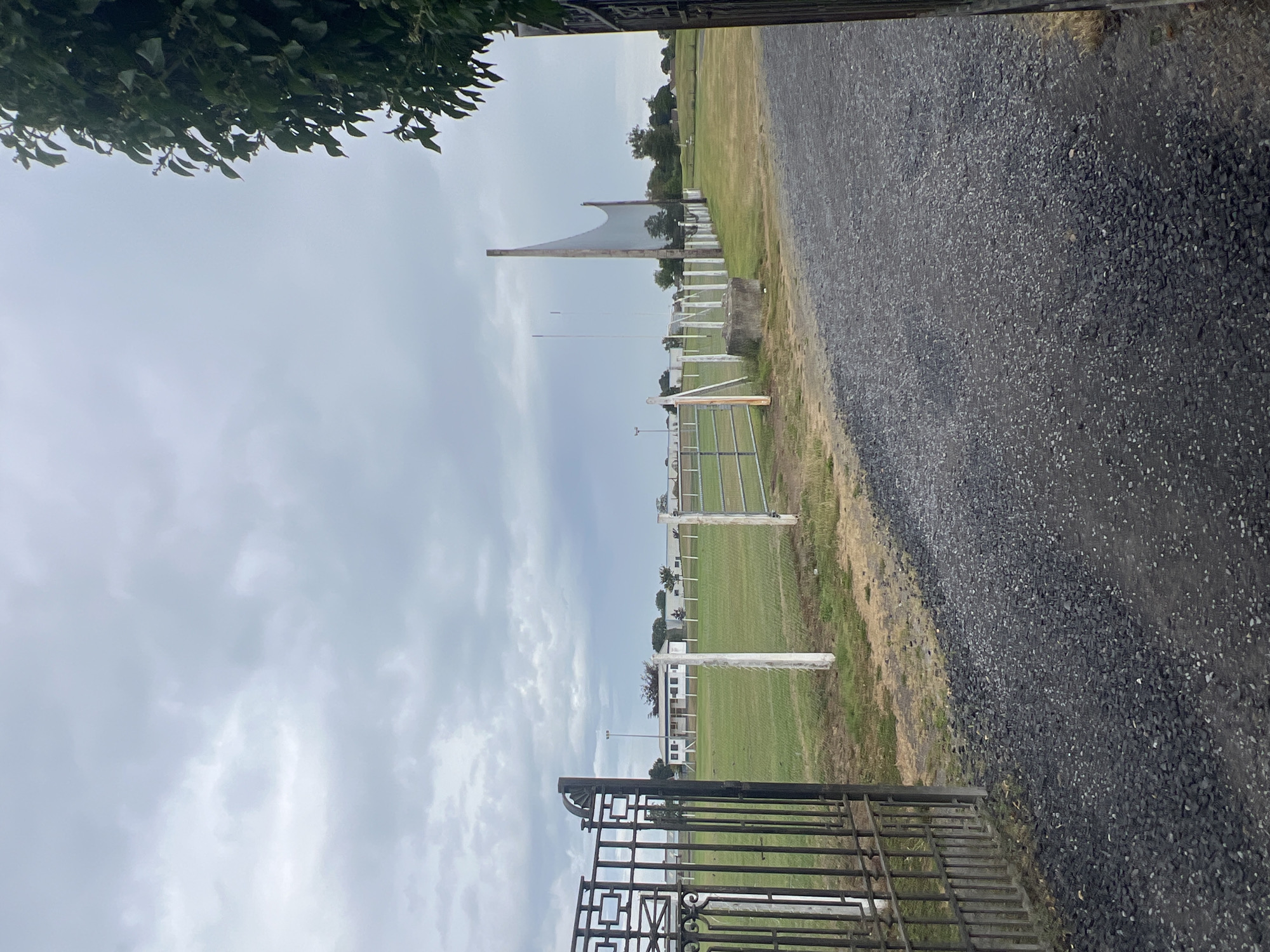 An open black metal gate stands on the left side of the frame, revealing a wide, gray, gravel driveway leading into an outdoor sports field. The field is covered in green grass and is surrounded by a wire fence. Several white posts support the fence, and some are capped with white cylinders. In the middle ground, a white building with a peaked roof and windows is visible. Beyond the building, several trees and smaller structures can be seen. On the right side of the field, a tall wooden post supports a large net, likely for sports like hurling or camogie. The sky is overcast with gray clouds.
