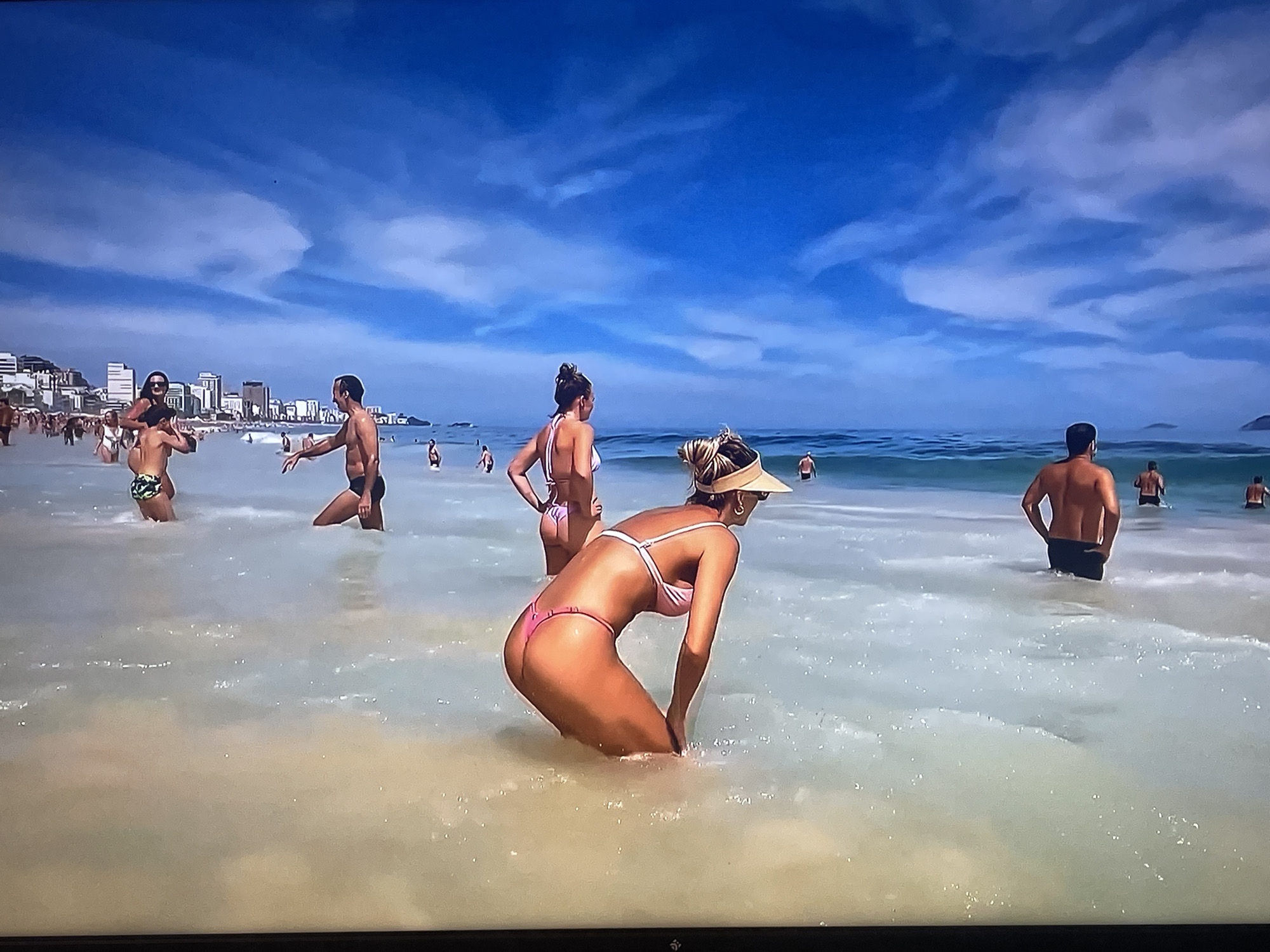 The image depicts a busy beach scene with several people in the water and on the shore. The foreground features a woman in a pink bikini bending down in shallow water. Further back, a woman in a white bikini stands, and a man in dark swim trunks is visible. On the left, a person in a patterned swim trunk is partially submerged. In the background, a city skyline with tall buildings is seen under a bright blue sky with scattered clouds. The water is clear and shallow, with gentle waves. The overall atmosphere is one of a lively summer day at the beach. The location is identified as the city of Shakhovskaya, Russia, although the visual elements do not specifically indicate this location and appear to be more characteristic of a coastal resort.