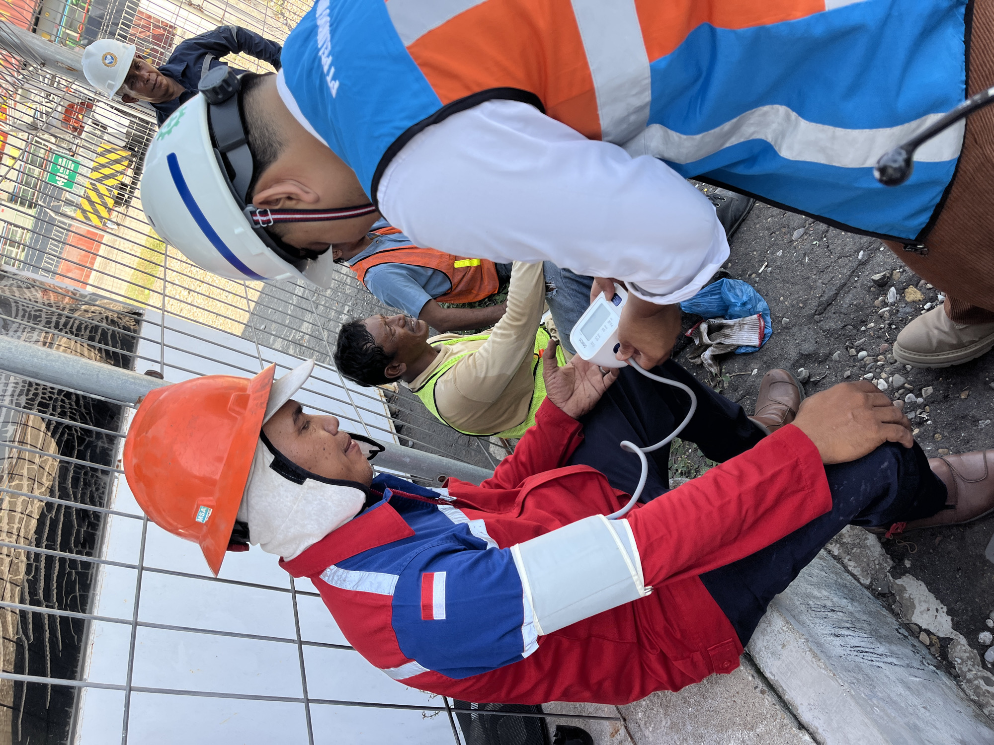 The scene is at Tambonbaroh, Indonesia. An injured man, wearing an orange hard hat, red and blue uniform, and brown shoes, is seated on the edge of a concrete surface near a metal fence. A medical professional in a white hard hat and blue and orange vest is taking the injured man's blood pressure using a digital monitor. Another individual in a yellow safety vest is seated nearby, looking on. The ground is a mix of asphalt and gravel.