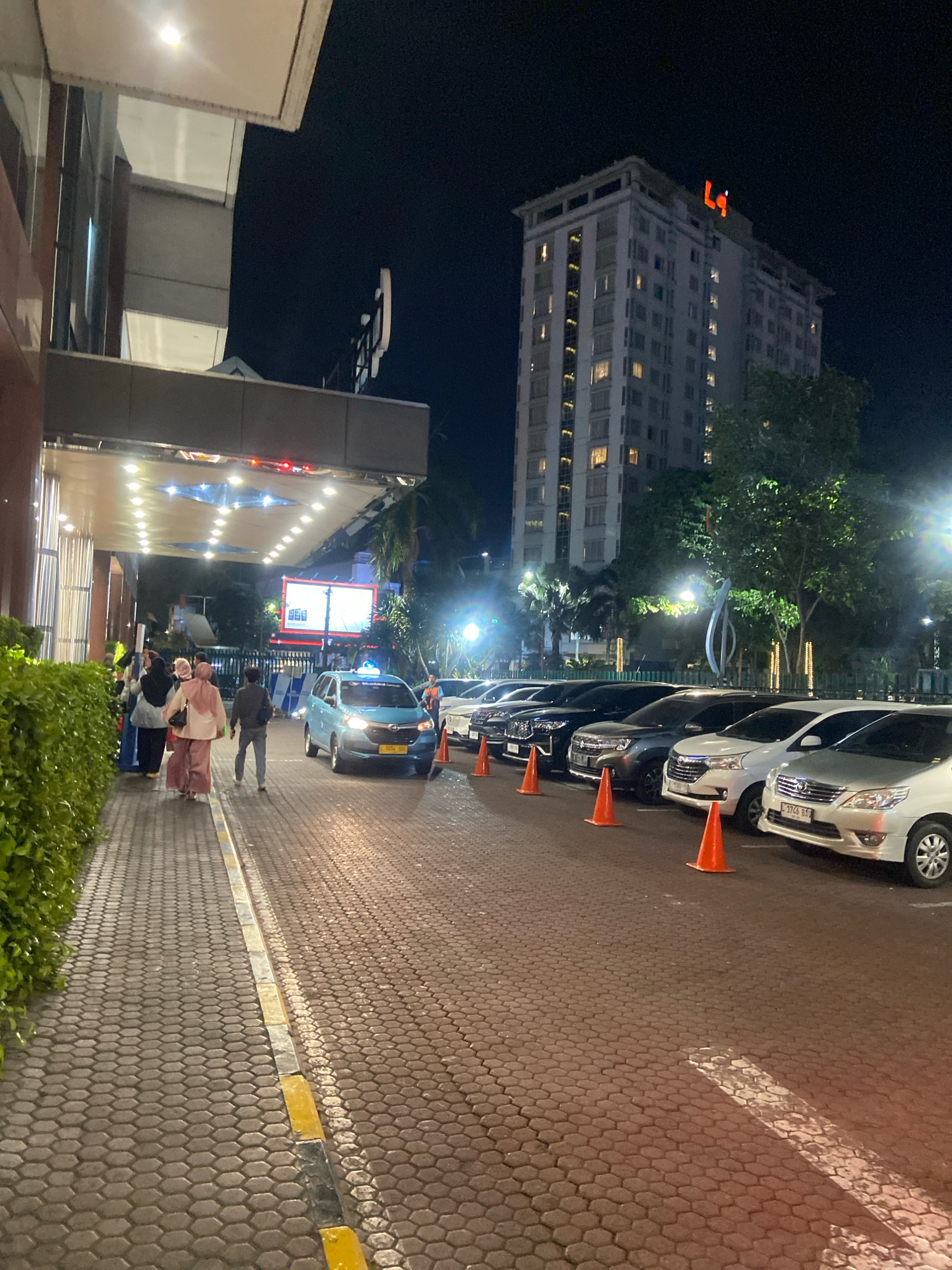 An exterior night shot shows a hotel or large building entrance and parking area in Surabaya, Indonesia. In the foreground, a paved walkway with hexagonal stones is on the left, bordered by a lush green hedge. A covered entrance area with bright lights is visible above the walkway, leading into the building. Several people, some wearing hijabs, walk on the walkway.
To the right of the walkway, a paved parking area is lined with several cars, including a light blue taxi with a visible license plate, and several white and dark-colored vehicles. Orange traffic cones are placed between some of the parked cars. In the background, a tall, multi-story building with many illuminated windows stands against the night sky. A large sign with orange letters, possibly a hotel logo, is visible at the top of this building. Trees and streetlights are also present in the background. The overall scene depicts a well-lit entrance and parking area of a commercial establishment at night.